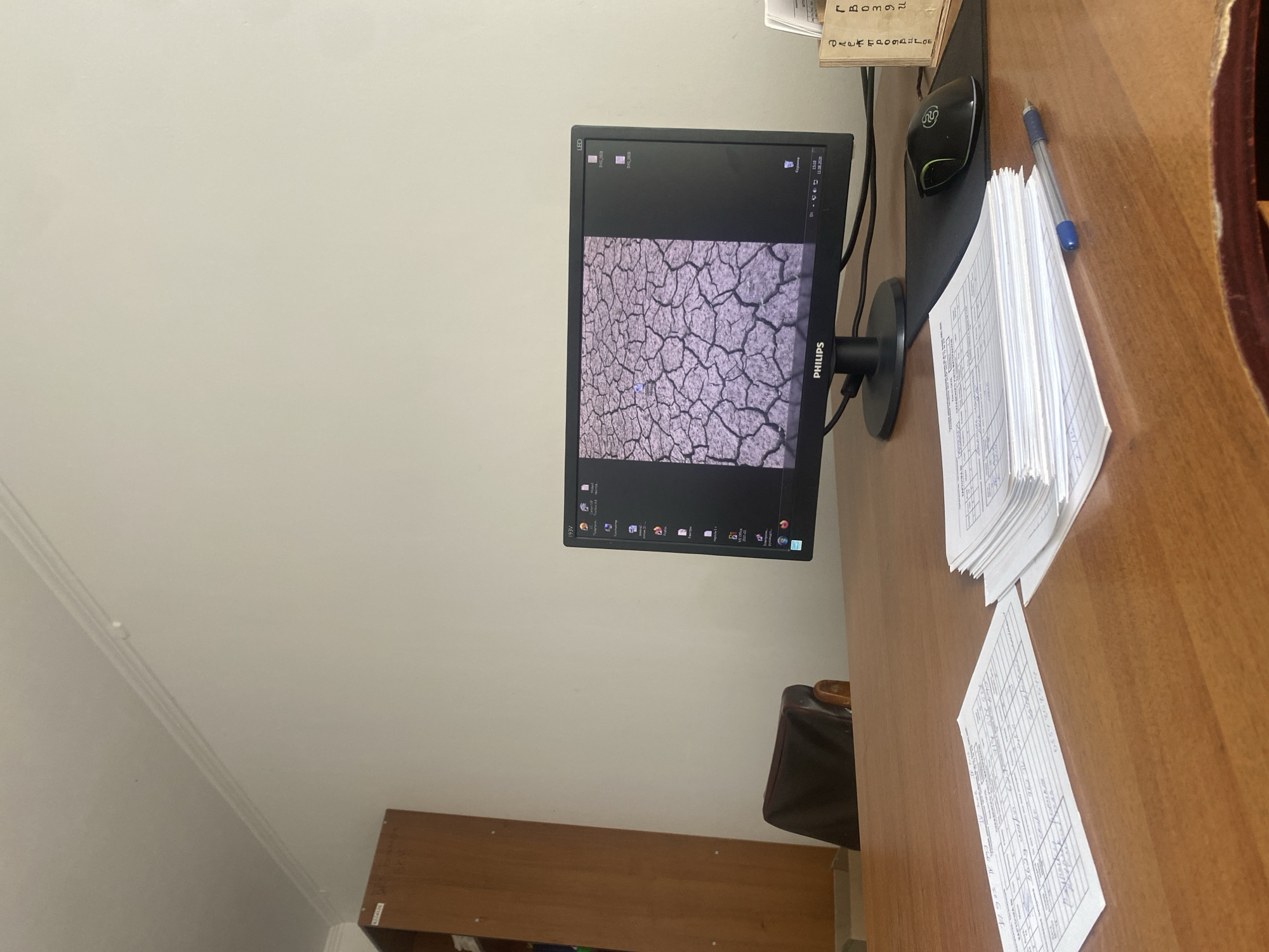 The image depicts an office desk with a computer monitor displaying a cracked earth wallpaper. A black Philips monitor is positioned centrally, with a black mouse to its right on a black mousepad. To the left of the monitor is a dark leather office chair, partially visible. In the foreground, a stack of white papers and a single sheet of paper with handwritten text are laid out on a wooden desk. A blue and silver pen lies on top of the papers. To the right of the monitor, a small wooden box with vertical lines of text is visible. Behind the desk, a tall wooden cabinet or bookshelf is positioned against a plain, light-colored wall. The ceiling is visible at the top of the image, featuring white molding. The scene is consistent with an office or workspace in Belovodskoe, Kyrgyzstan.