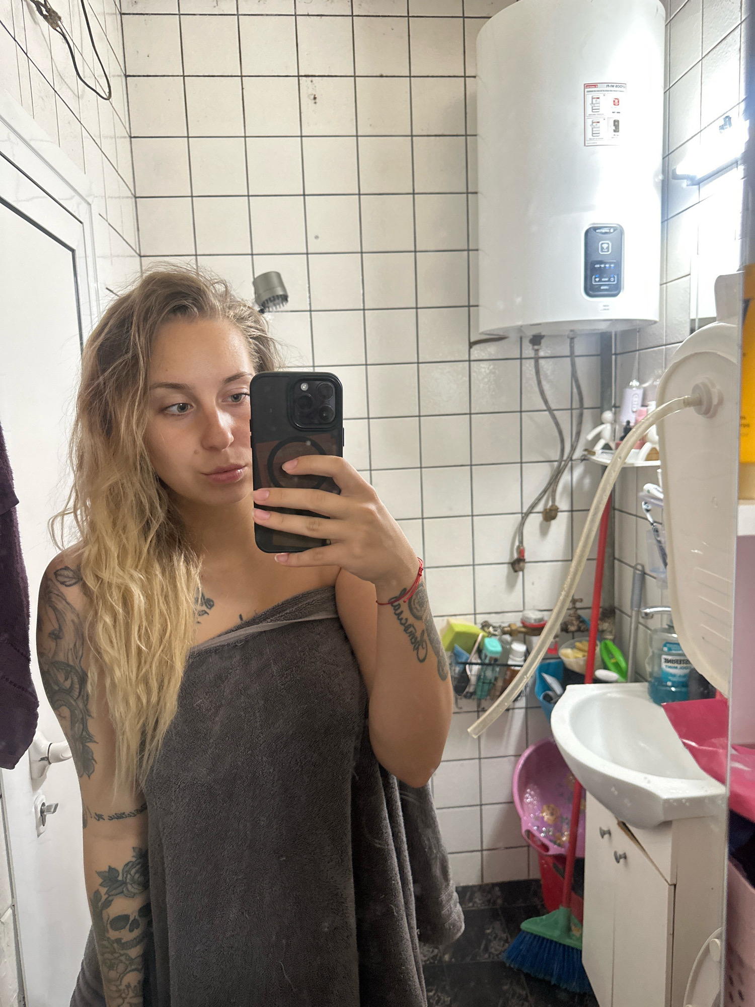 A young woman with blonde hair, wearing a grey towel, takes a mirror selfie in a bathroom. She has several tattoos on her arm. The bathroom walls are tiled with white square tiles, and a white water heater is mounted on the wall. There is a sink, a shower head, and various cleaning supplies visible. The floor is black and white checkered.
