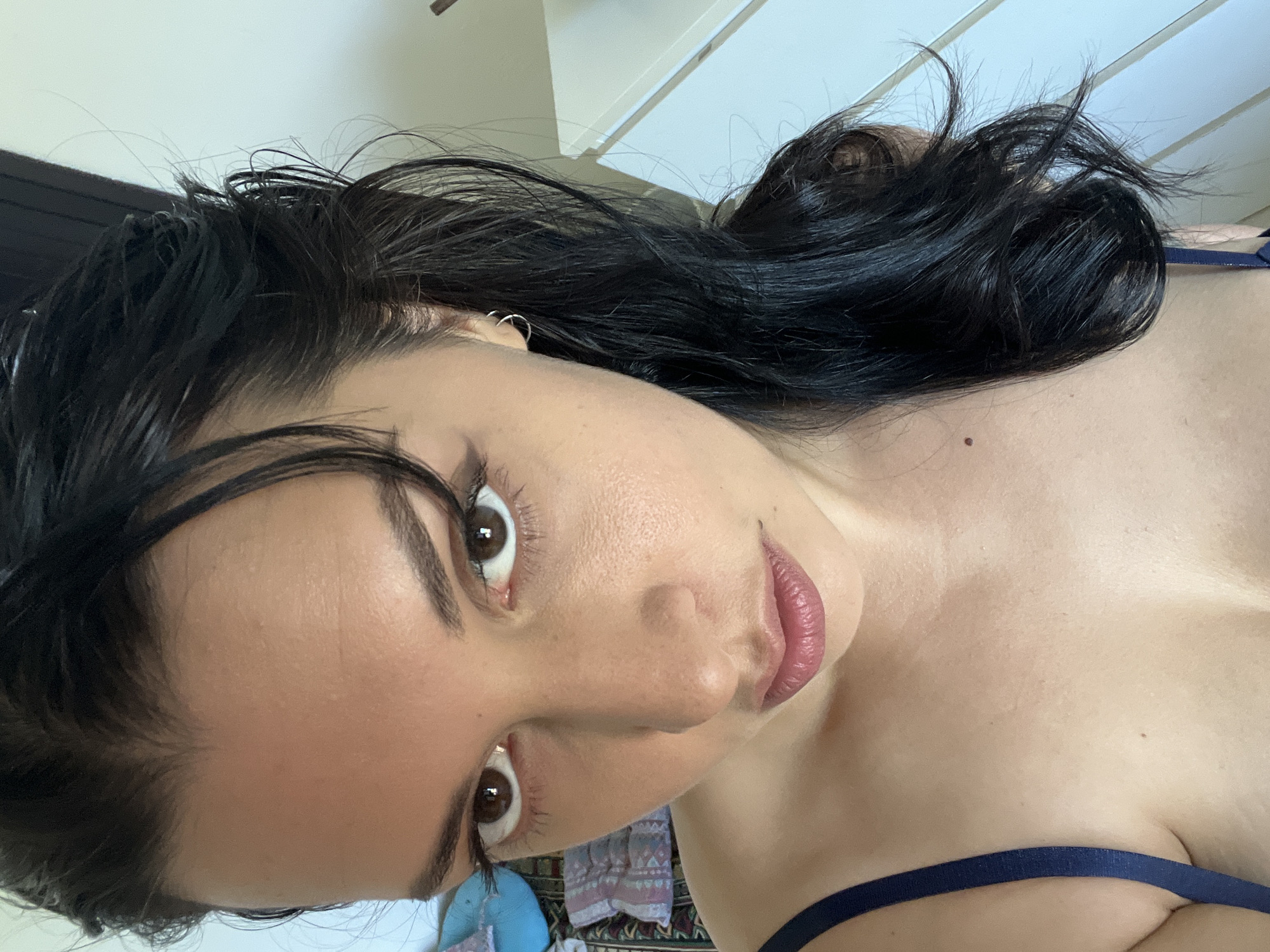 A close-up, high-angle selfie of a woman with dark hair and brown eyes. She is looking directly at the camera with a serious expression. She is wearing a dark blue bra strap visible on her left shoulder. Her hair is dark and wavy, partially obscuring her face on the right side. The background is out of focus, but suggests an indoor setting with hints of furniture and patterned fabric. There is no indication of the city of Đà Nẵng, Vietnam in the image.