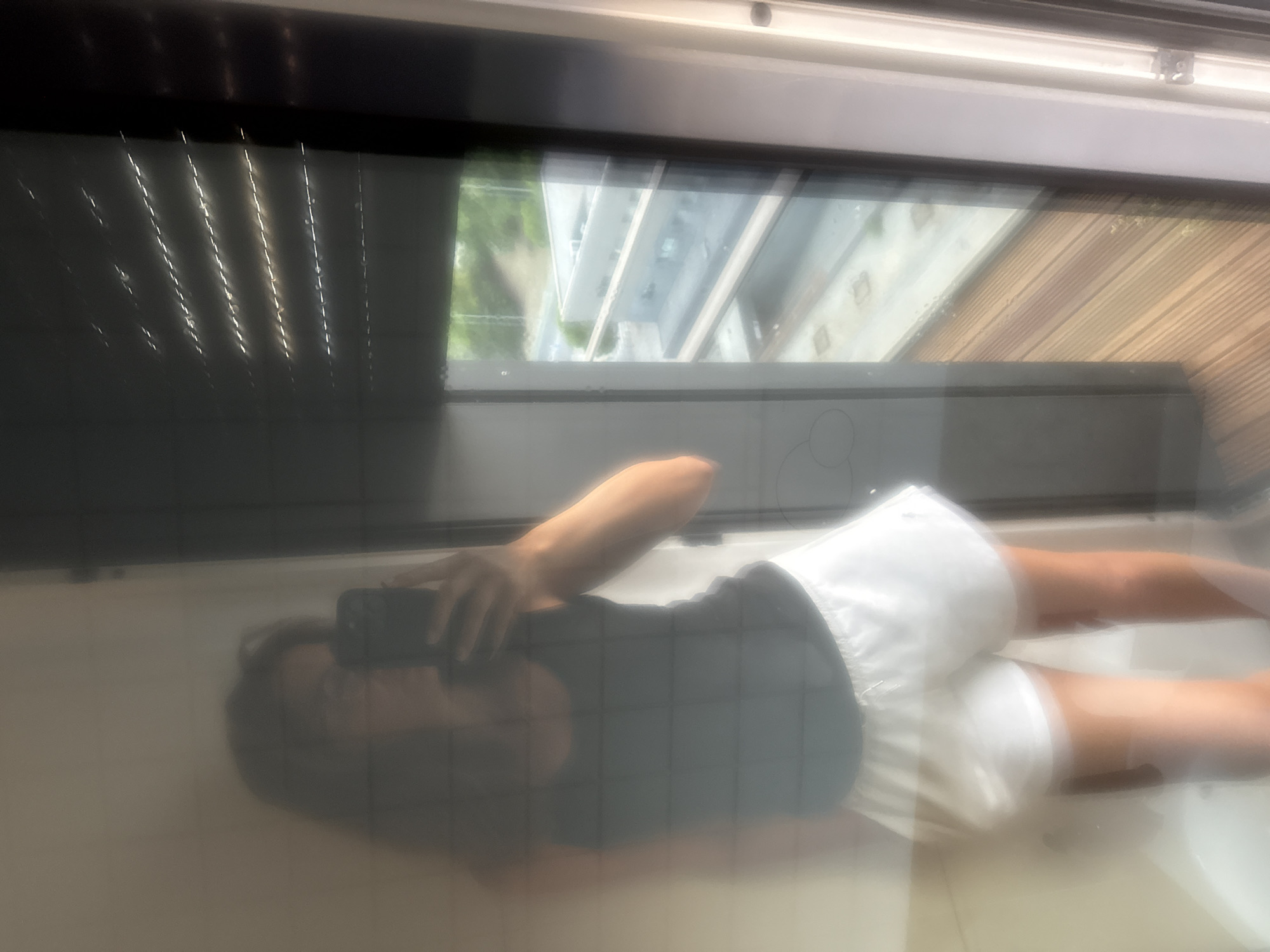 This media file shows a reflection of a person, likely taken from a smartphone. The person is wearing a black tank top and white shorts. They are holding a smartphone in their right hand, as if taking a selfie. The reflection is distorted, possibly due to the surface it's reflecting off, which appears to be a window or glass surface.
Through the window, an outdoor scene is visible, including buildings and trees, suggesting an urban or suburban environment. The lighting suggests it might be daytime. The image is somewhat blurry, with a noticeable grid pattern overlaying the person's reflection, which could be a visual artifact from the capturing device or the surface itself.
The scene is likely set indoors, near a window. There are blinds visible on the left side of the frame, partially obscuring the view outside. The overall impression is a casual, personal moment captured in a reflective surface. There is no visible text in the image.