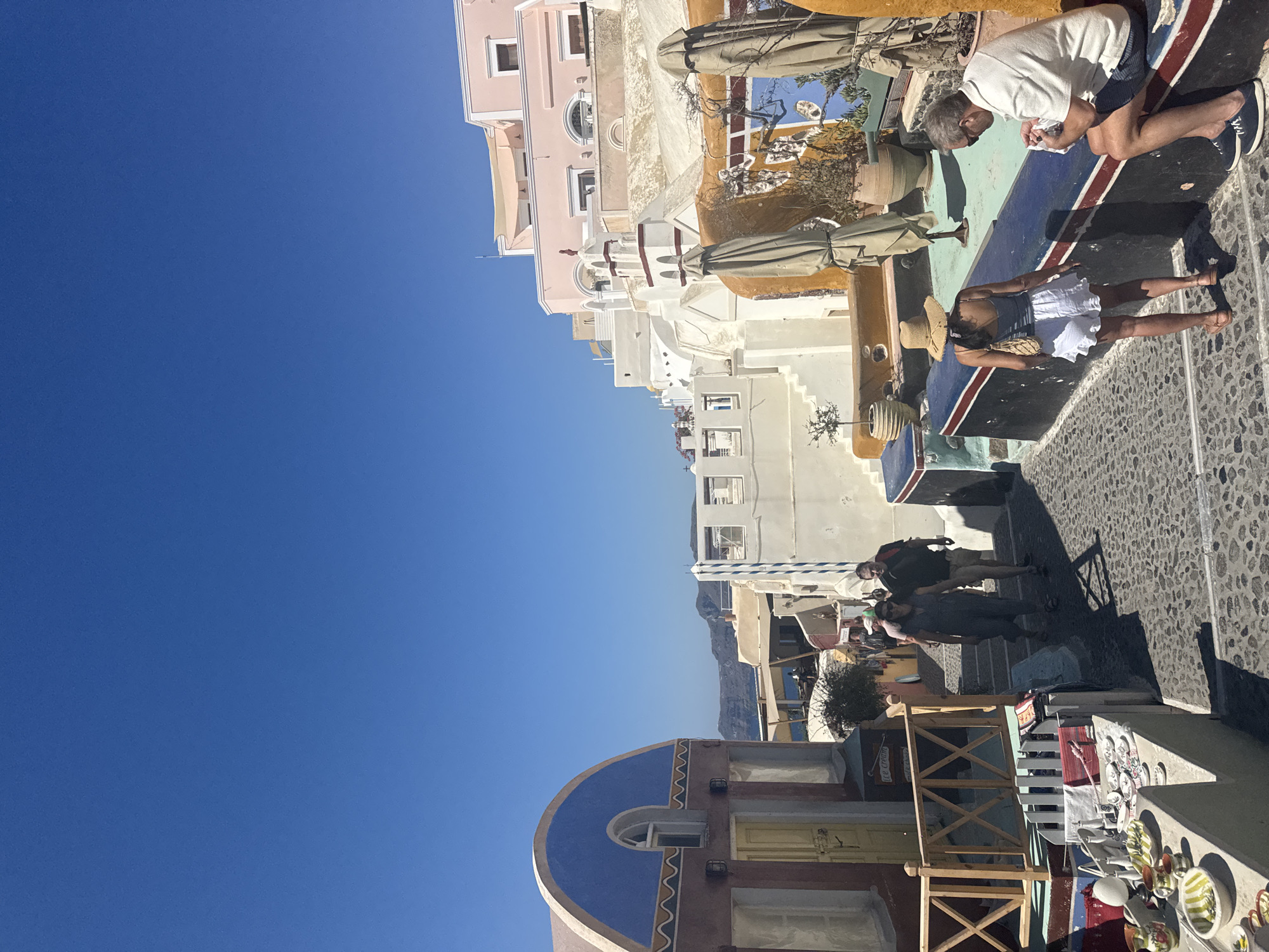 The image depicts a sunny day in Oia, a municipal unit of Santorini, Greece. The scene captures a narrow cobblestone street lined with traditional white-washed buildings, characteristic of the Cycladic architecture. The buildings feature colorful accents, including a prominent blue arched roof on the left and pink structures on the right.
In the foreground, to the left, a shop displays various ceramics and souvenirs on a low wall. Further up the street, several people are walking. A couple, a man and a woman, are seen walking away from the camera. The woman wears a grey dress and sunglasses, while the man wears a black t-shirt and shorts.
To the right, a woman wearing a straw hat and white shorts is walking towards the left, facing away from the camera. A man is seated on a low wall on the right, wearing a white polo shirt and blue shorts. He appears to be looking at something in his hands.
Several closed umbrellas are visible, suggesting a cafe or seating area. In the background, more buildings ascend the hillside, with glimpses of the Aegean Sea and distant islands under a clear blue sky. The overall impression is one of a bustling tourist destination in a picturesque Mediterranean setting.