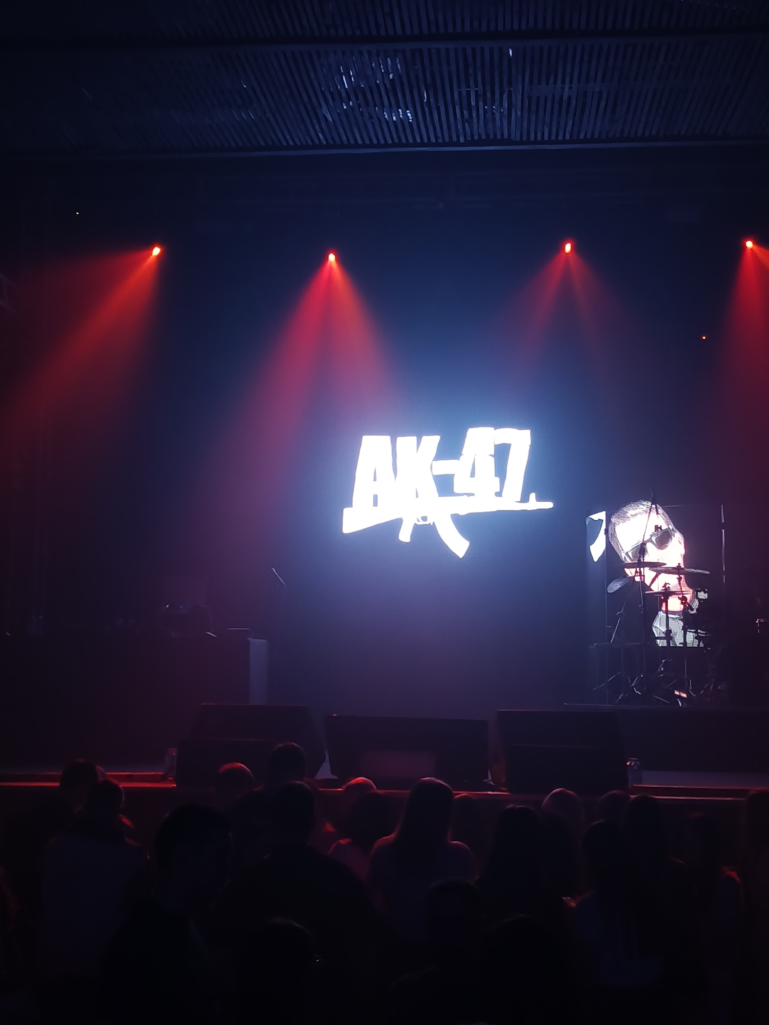 Here's a forensic description of the image:
**Scene:** The image depicts a concert or performance setting. The location is likely a venue in Izhevsk, Russia, given the presence of "AK-47" as a prominent symbol on the stage, which may be a reference to the city's history as a major manufacturer of the firearm.
**Environment:** The environment appears to be dimly lit, with red stage lighting casting beams down onto the performance area. The ceiling has a slatted design.
**Subjects:** There is a crowd of people in the foreground, viewed from the back. Their faces are not clearly visible due to the low lighting.
**Objects:**
* A large screen displays the text "AK-47" with an image of the rifle incorporated into the logo. To the right of the logo, there's a projected image that includes a drummer playing.
* Speakers are positioned on the stage in front of the screen.
* Musical equipment, potentially a drum set, is seen on the stage behind the screen.
**Actions/Interactions:** The people in the crowd are likely attending the concert and watching the performance. The performer on the screen is playing the drums.