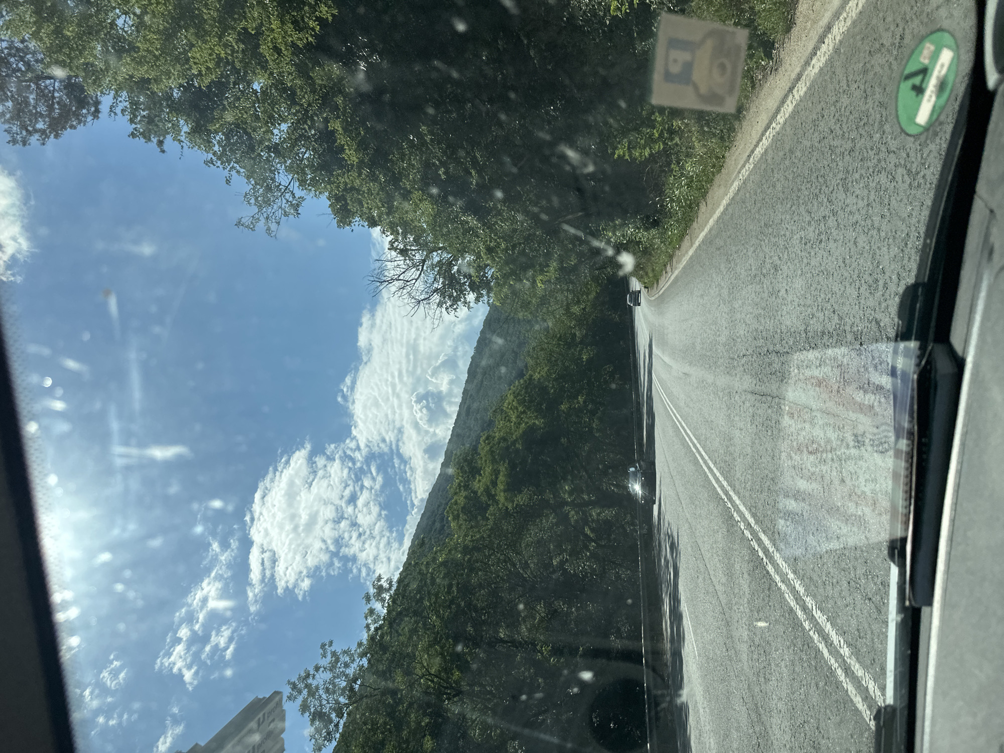 The image depicts a view from a car driving on a paved road through a forested, mountainous area. The road has double white lines in the center. Lush green trees line the right side of the road, and the dense foliage suggests a natural, possibly rural, environment. On the left side of the frame, a bright blue sky with scattered white clouds is visible. The sun is shining, casting shadows on the road, indicating it is daytime. There are two stickers on the inside of the windshield. One is a square sticker with a yellow bear-like figure and a blue background. The other is a circular green sticker with white text and symbols, possibly a vehicle inspection or environmental sticker. A small dark-colored car is visible ahead on the road. The scene conveys a sense of travel and exploration through a scenic, natural landscape. The image is oriented vertically, with the road running from the top left to the bottom right.
