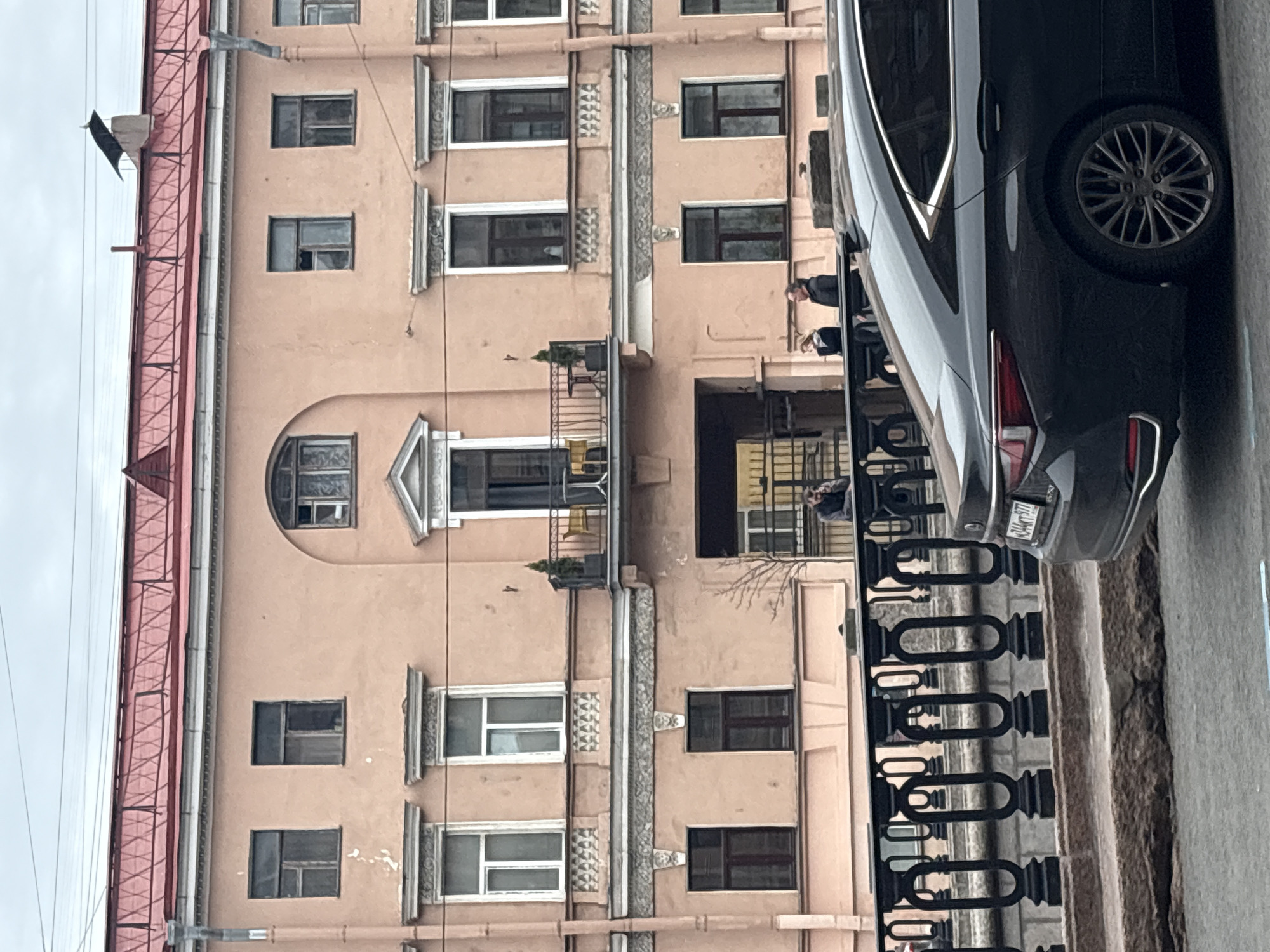 The photograph captures a building facade in Saint Petersburg, Russia. The building is painted in a light peach color and has multiple windows arranged symmetrically. Architectural details are present around the windows, adding to the building's character. A balcony is visible on one of the floors. The photograph also shows a portion of a black car parked on the side of the building with the license plate covered. A decorative black fence is running along the street. People can be seen on the balcony. The weather appears overcast. The image has been rotated.