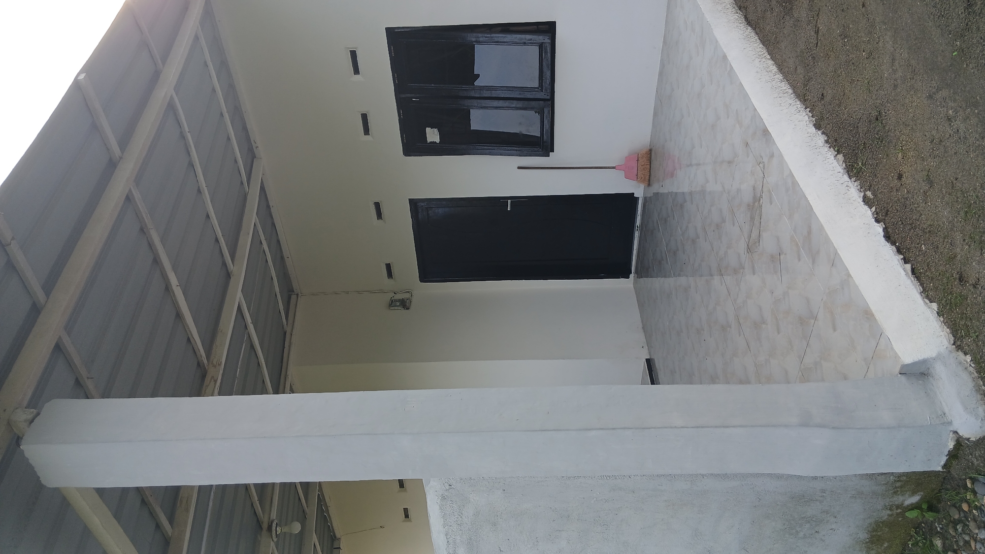 The image depicts the exterior of a house located in Klambir Lima Kebun, Indonesia. A large, white concrete pillar stands to the left, supporting a corrugated metal roof structure. Adjacent to the pillar is a textured, light gray wall. The main structure of the house is painted white, featuring a black door and a black-framed window with two panes. A pink broom leans against the wall near the window. The ground in front of the house is covered with light-colored, square tiles, and a white concrete border runs along the edge. The area outside this border is a mix of dirt and sparse vegetation. The overall scene suggests an outdoor area adjacent to a residential building, possibly a porch or veranda.