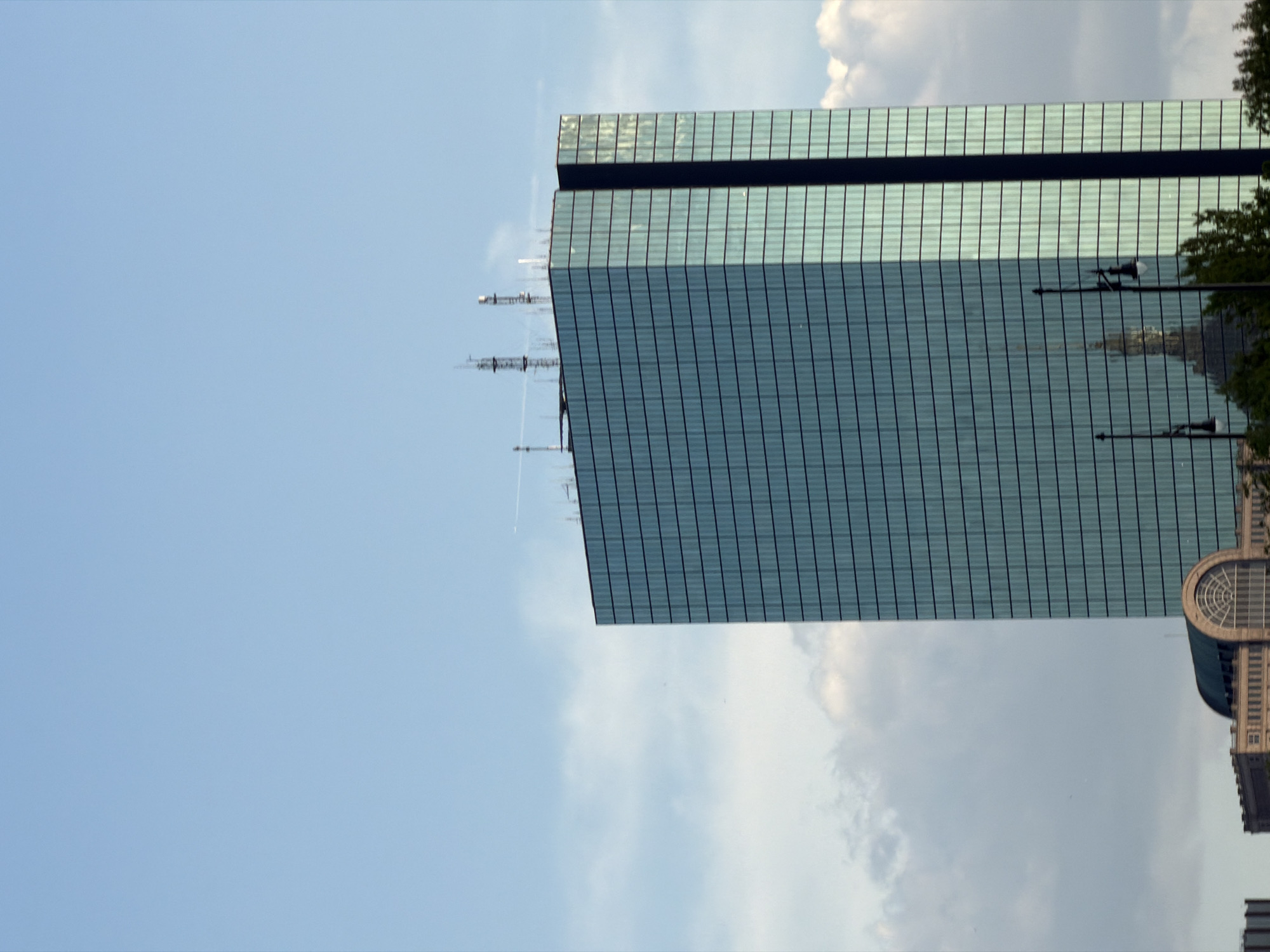 The image depicts the John Hancock Tower in Boston, Massachusetts. The blue glass-clad skyscraper dominates the view, with a clear sky and fluffy white clouds in the background. Antennas are visible on the top of the tower. A portion of another building, possibly the Boston Public Library, is reflected in the glass facade of the John Hancock Tower. A streetlight is also visible in the reflection.