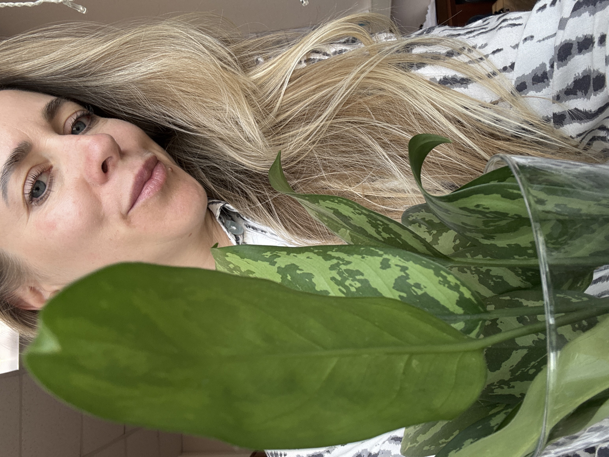 A Caucasian woman with blonde hair is holding a plant in a glass vase. She is wearing a patterned shirt. The exact location in Chaikovsky, Russia, cannot be determined from the image.