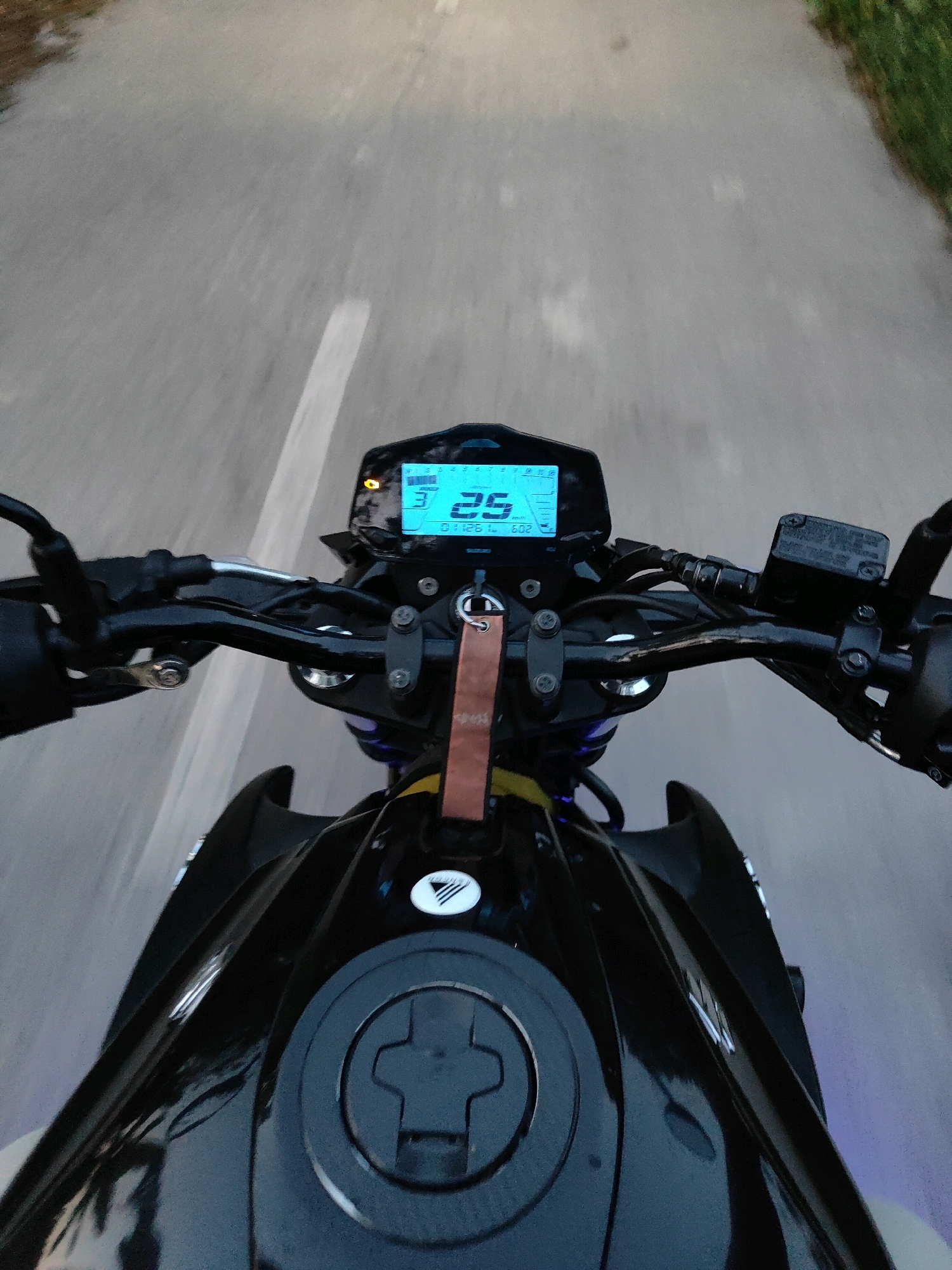 The image is a first-person perspective shot from a motorcycle, likely in Senbagh, Bangladesh, as the road surface and surrounding greenery are consistent with rural or semi-urban environments in the region. The motorcycle is black and the rider is in motion, indicated by the blurred asphalt road with a white lane marking. The motorcycle's digital dashboard is visible, displaying a speed of 25 km/h. The handlebars, brake fluid reservoir, and part of the fuel tank are also in view. A small, brown tag hangs from the handlebars. The lighting suggests it might be dusk or dawn. There are no people or other vehicles clearly visible in the frame.