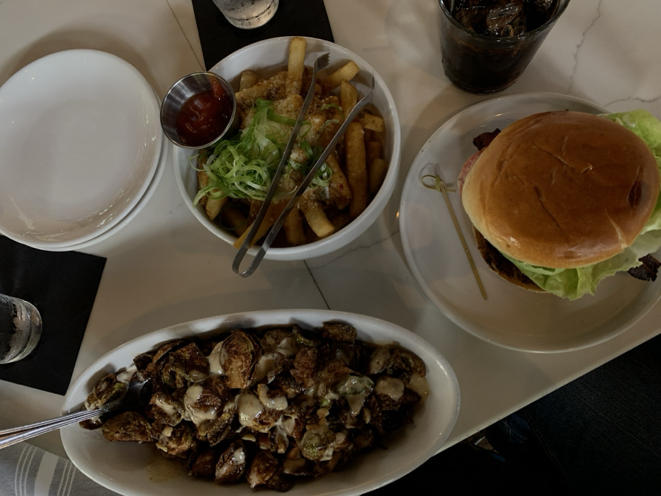 An overhead image captures a dining setup on a light-colored, reflective table surface in Osage Beach, United States. The scene features three main food presentations. A white oblong dish, positioned on the left, contains roasted Brussels sprouts extensively drizzled with a light-colored, creamy sauce. A silver serving spoon is resting within this dish. To its right, a white round bowl holds French fries topped with sliced green onions and a visible granular seasoning. A small metal ramekin containing red ketchup is placed on the edge of the fry bowl, and metal serving tongs are resting across the fries. In the foreground, a white round plate displays a hamburger with a brioche bun, lettuce, and visible bacon; a small wooden skewer lies on the plate. A dark beverage with ice is contained within a textured dark glass to the right of the hamburger. A clear glass of water is partially visible at the top of the frame. A dark fabric napkin is positioned beneath the water glass, and a striped fabric napkin is partially visible on the left side of the table.