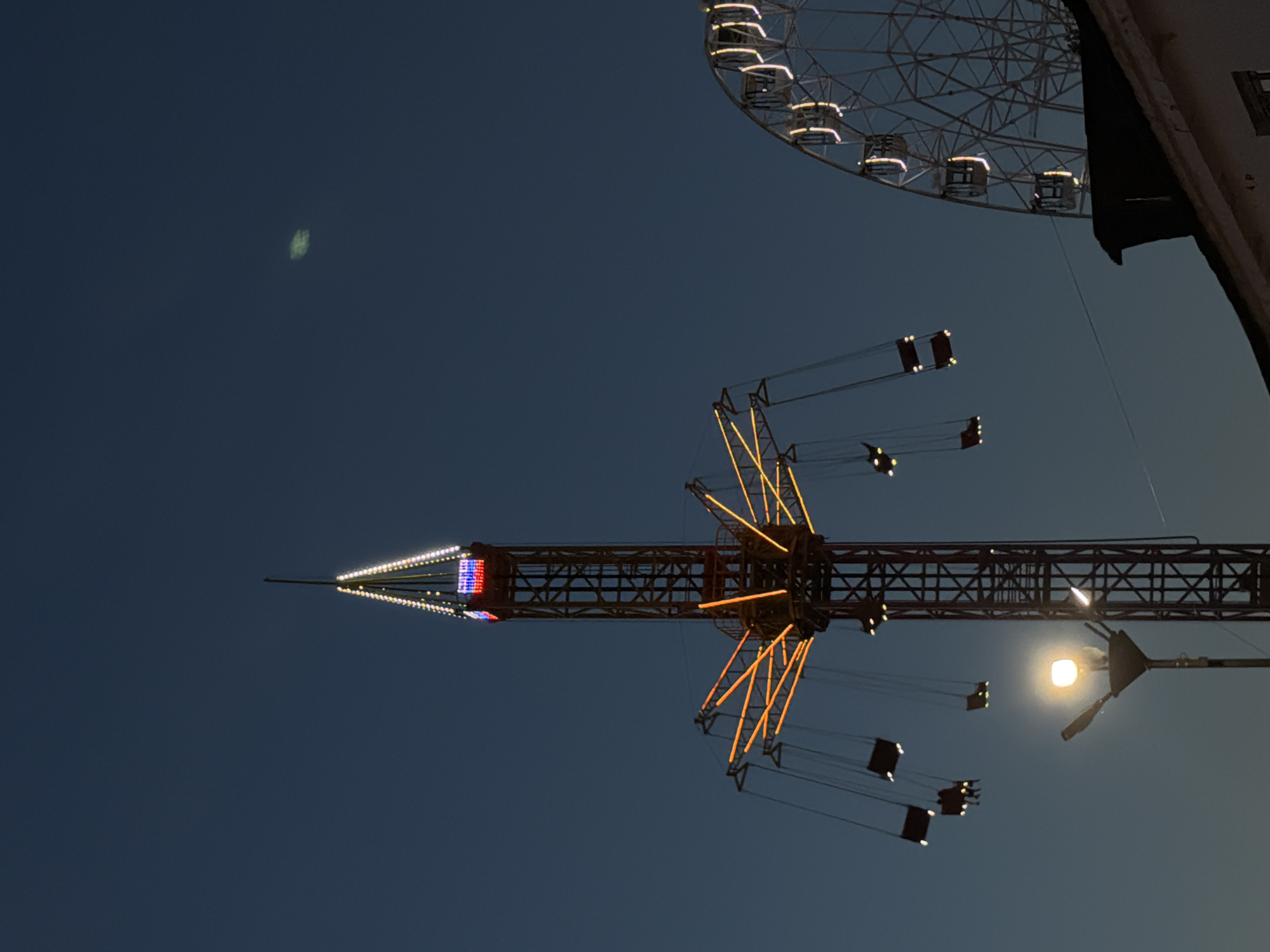 The image shows amusement park rides against a dark blue sky. A swing ride with illuminated arms and seats occupies the lower center. The ride has a central tower decorated with red, white, and blue lights. A portion of a Ferris wheel with lit-up passenger cars is visible in the upper right corner. A faint, blurred object is visible in the upper left quadrant of the sky. The photograph appears to be taken at night or during twilight. The location is Izhevsk, Russia.