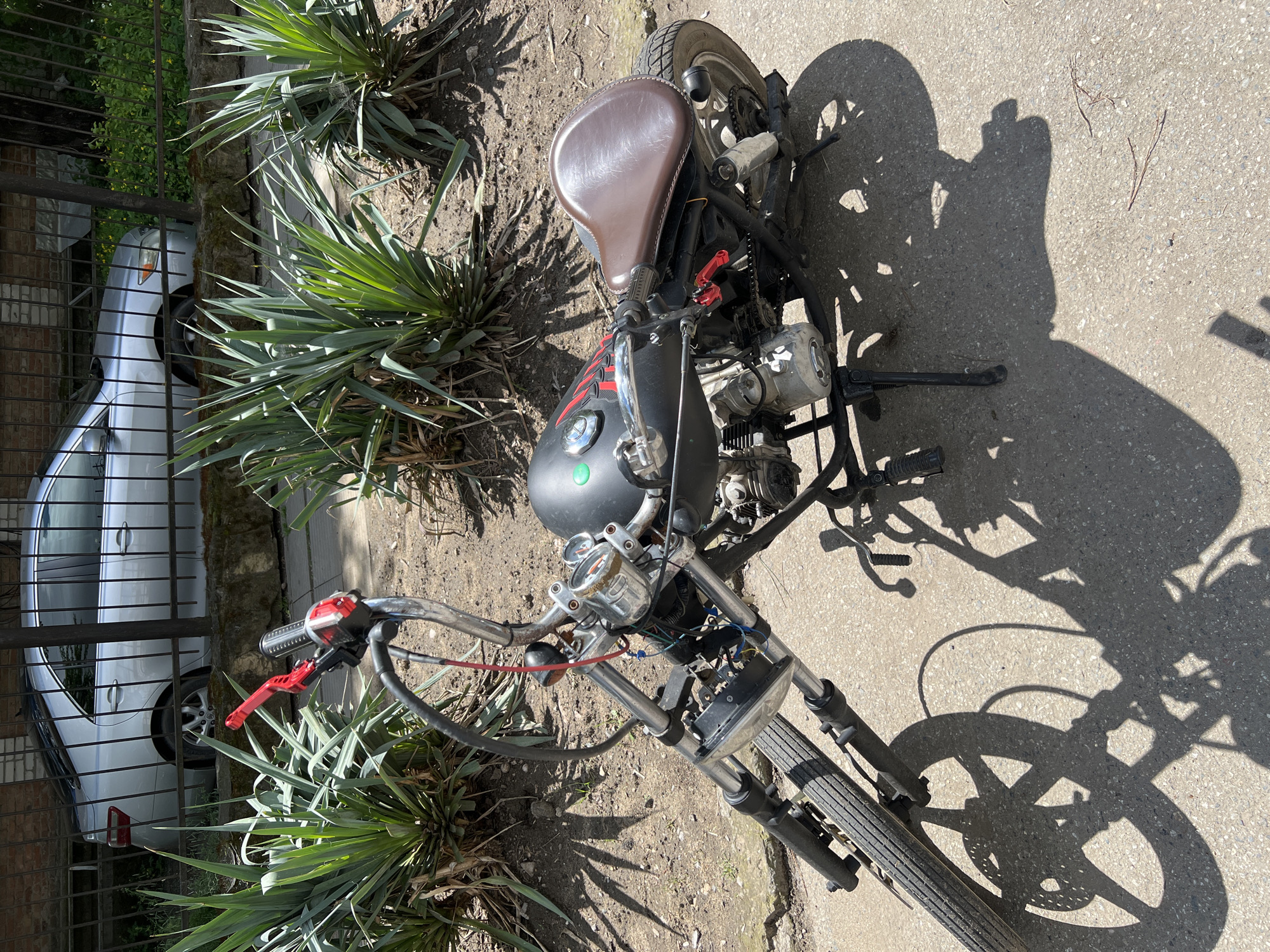 The image shows a motorcycle parked on a concrete surface next to a patch of greenery. The motorcycle has a brown leather seat and a black fuel tank. A silver car is visible behind a fence. The photo was taken in Krasnodar, Russia.