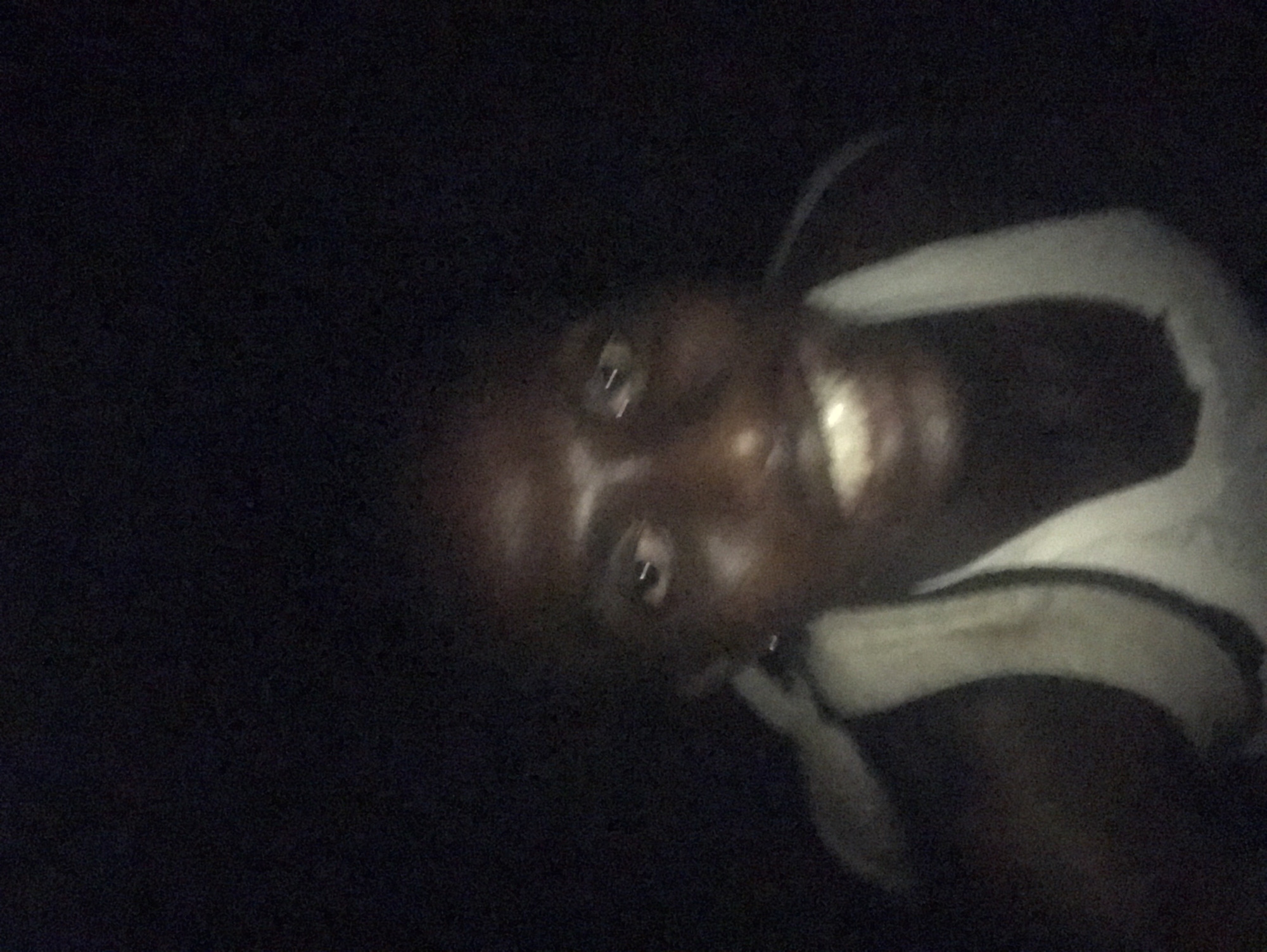 This is a low-light, grainy photograph of a person with dark skin, captured in a very dark environment, likely at night. The image is rotated sideways. The person's face is partially illuminated, showing both eyes and a broad smile that reveals their teeth. Light reflects in their eyes, suggesting a direct light source, possibly a camera flash or screen. They are wearing a light-colored, possibly white, sleeveless garment with straps visible over their shoulders. The background is uniformly dark, providing no discernible details about the setting or location. The person is looking directly at the camera, implying a selfie or direct interaction with the photographer. Their smile suggests an emotion of happiness or amusement. No specific objects or text are visible besides the person and their clothing.