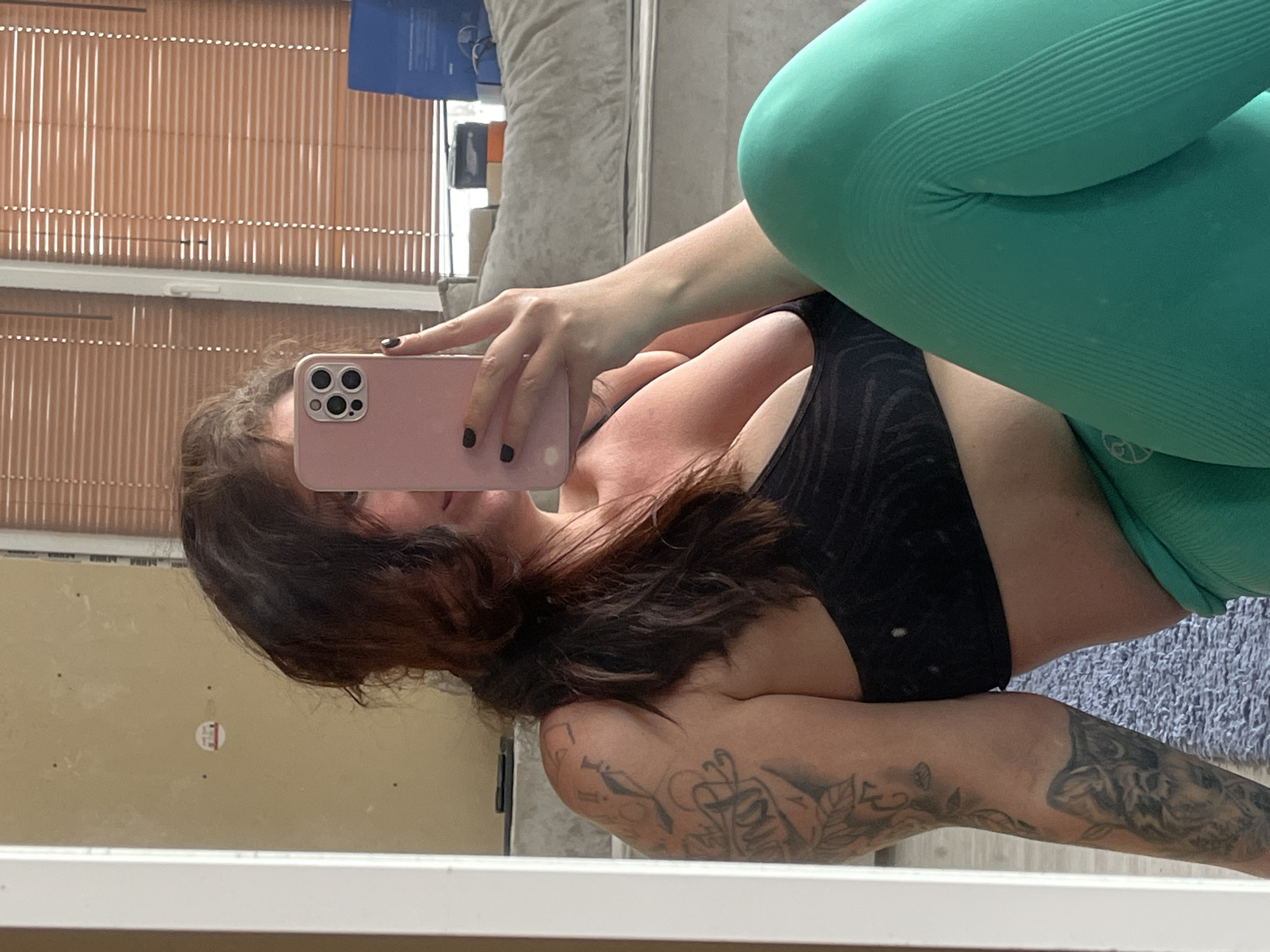 A woman with dark hair is taking a selfie in a mirror. She is wearing a black sports bra and green leggings. Her left arm is visible and has multiple tattoos. The background includes a window with blinds, a wall, and a portion of a couch. The location is indicated as Мосметаллоконструкция Онт, Russia, but there is no information in the image to confirm this.