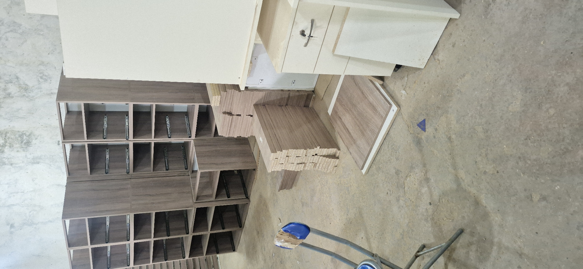 The image shows a furniture assembly area. A partially assembled brown cabinet with multiple shelves is positioned on the left side. Adjacent to the cabinet are stacks of brown wood panels, possibly components for additional shelves or cabinets. Above these, a white cabinet or drawer unit is visible. A blue triangle marker is on the floor in the lower right quadrant. The scene is indoors, possibly a workshop or storage area.