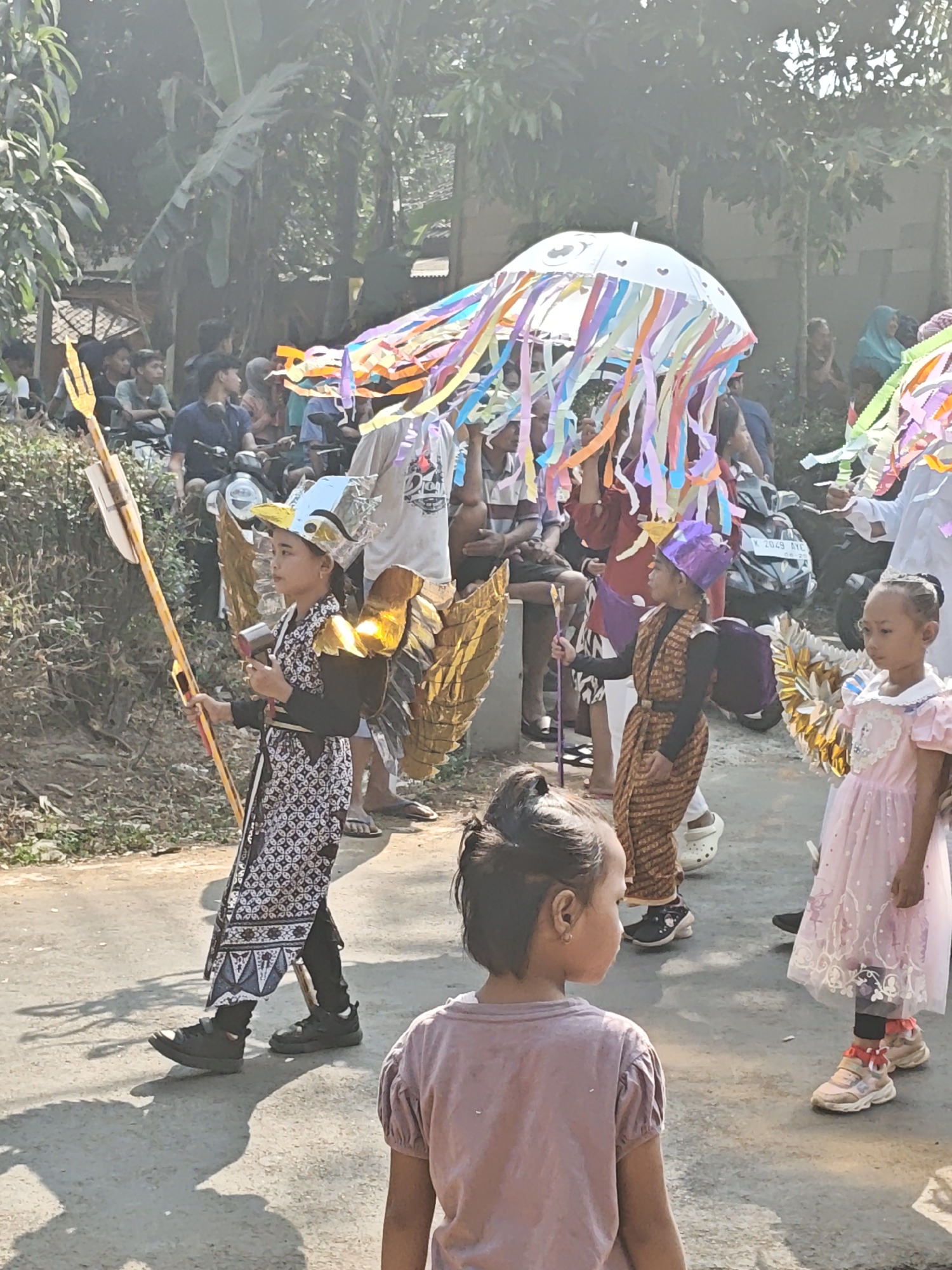 In Ngasem, Indonesia, children participate in a festive parade, likely a cultural event. In the foreground, a young girl with her back to the camera wears a light purple t-shirt. To her right, a girl in a pink dress with ruffled sleeves and white accents stands looking to her left. Behind her, a child in a brown and gold patterned outfit with a purple headdress holds a staff. To the left, a girl in a black and white patterned robe and black pants holds a large golden trident and wears golden wings. She also has a gold and silver headdress. In the background, spectators watch the parade, many seated on motorcycles. A large white umbrella decorated with colorful streamers, resembling a jellyfish, is held aloft. The event takes place on a paved road with a dirt shoulder, lined with trees and vegetation.