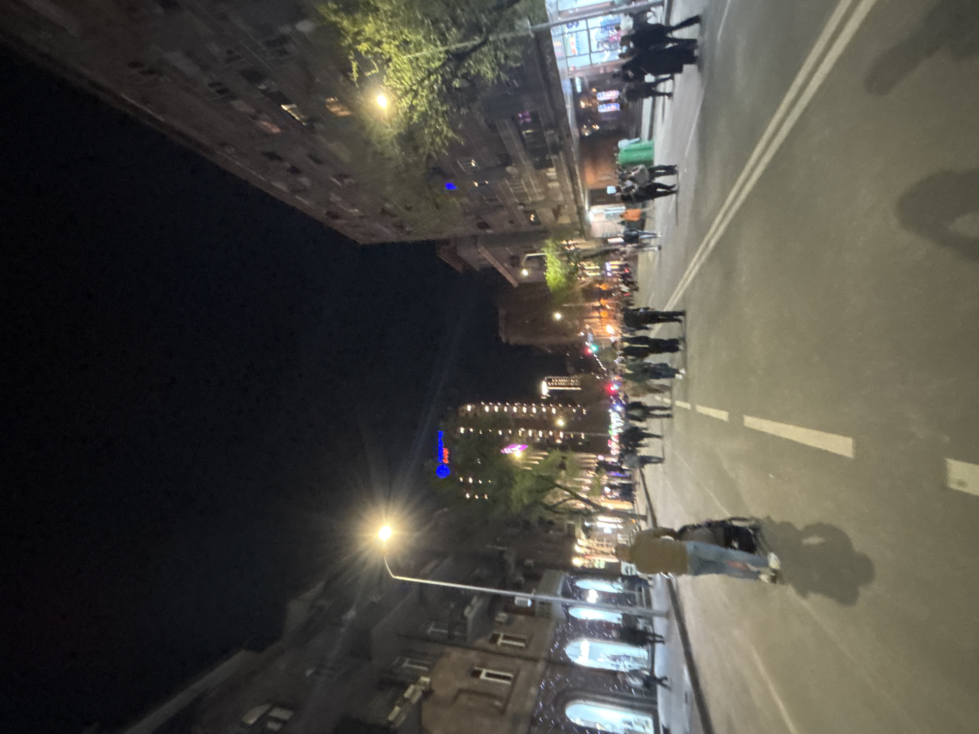 The image captures a vibrant urban street scene at night in Yerevan, Armenia. The view looks down a well-lit street flanked by multi-story buildings and trees.
Numerous people are present, walking and standing on both the pavements and what appears to be a road surface marked by a double white line. Their figures are visible in silhouette and partial illumination, but individual details are obscured by the low light and distance. The overall activity suggests a casual evening gathering or promenade.
The scene is illuminated by artificial light sources. Prominent streetlights cast bright light, with one in the foreground creating a starburst effect and long shadows of people on the ground. Buildings along the street feature lit windows and some are adorned with string lights, enhancing the festive night ambiance. Trees with green foliage are also visible, catching the light. A distinct blue glowing sign or decoration can be seen on a building further down the street, and a green bin or container is visible on the pavement. The architecture of the buildings appears to be multi-storied and possibly historic. No legible text is discernible within the image.