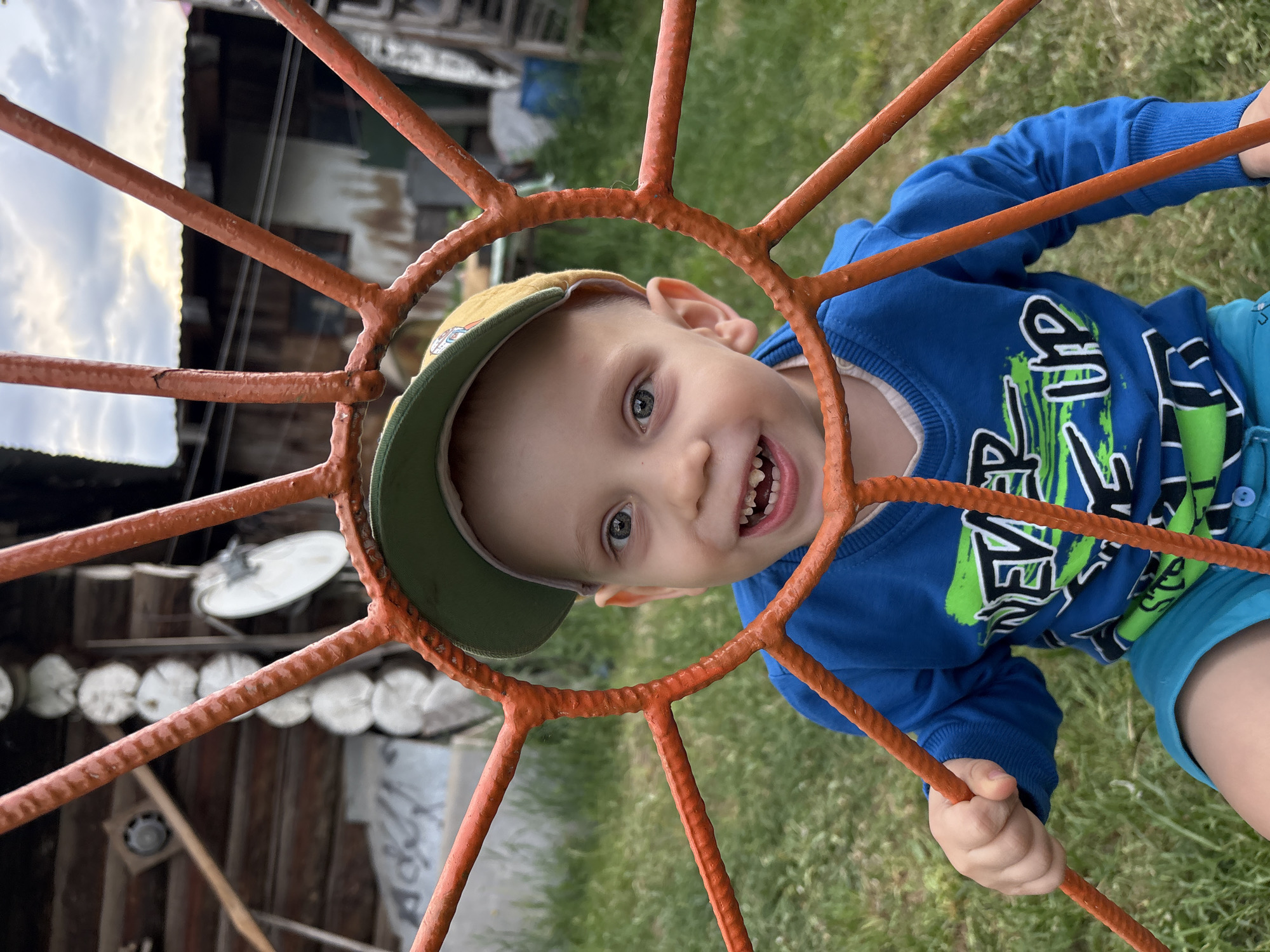 A young boy is seen looking through a circular, orange-painted metal structure with radiating spokes. He is wearing a blue long-sleeved shirt with green graphics, blue shorts, and a yellow cap with a green brim. He is smiling, revealing his teeth. The background shows a grassy area and a structure made of logs, as well as a satellite dish. The location is Chaikovsky, Russia.