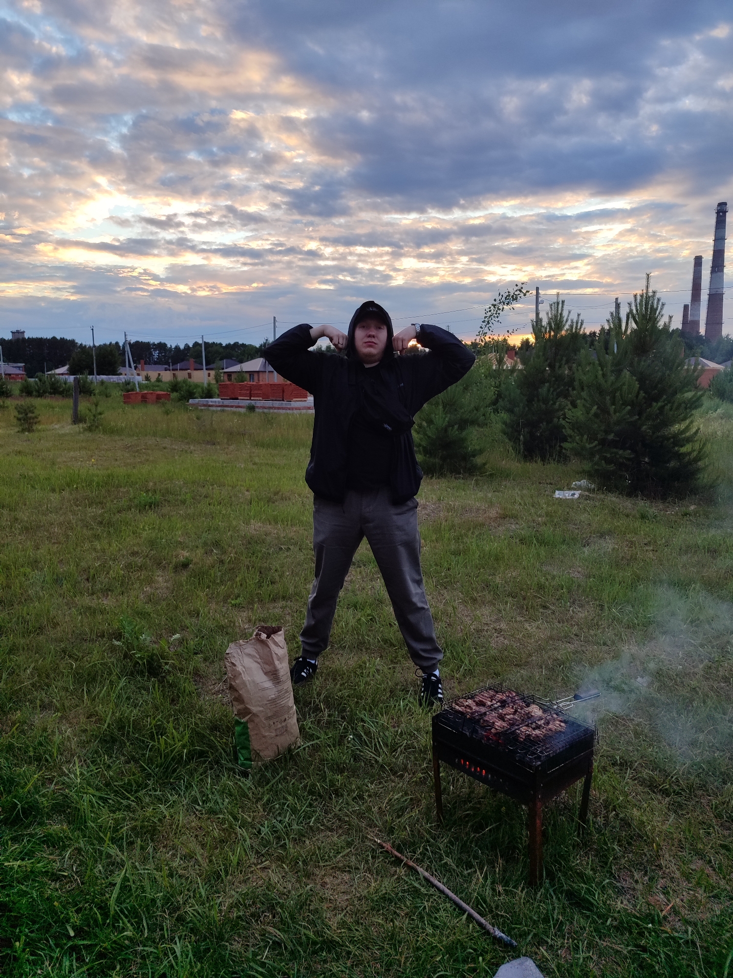 A young man stands in a grassy field in Chaikovsky, Russia, flexing his biceps under a dramatic, cloudy sky. He wears a black hoodie and gray pants, with a black bag slung across his chest. In the foreground, a charcoal grill sizzles with meat, emitting a thin wisp of smoke. To the left of the grill, a brown paper bag, likely filled with charcoal, rests on the grass. In the background, several pine trees stand tall, and beyond them, hints of residential buildings and industrial smokestacks suggest the edge of a settlement. The overall scene captures a casual outdoor gathering with a barbecue.