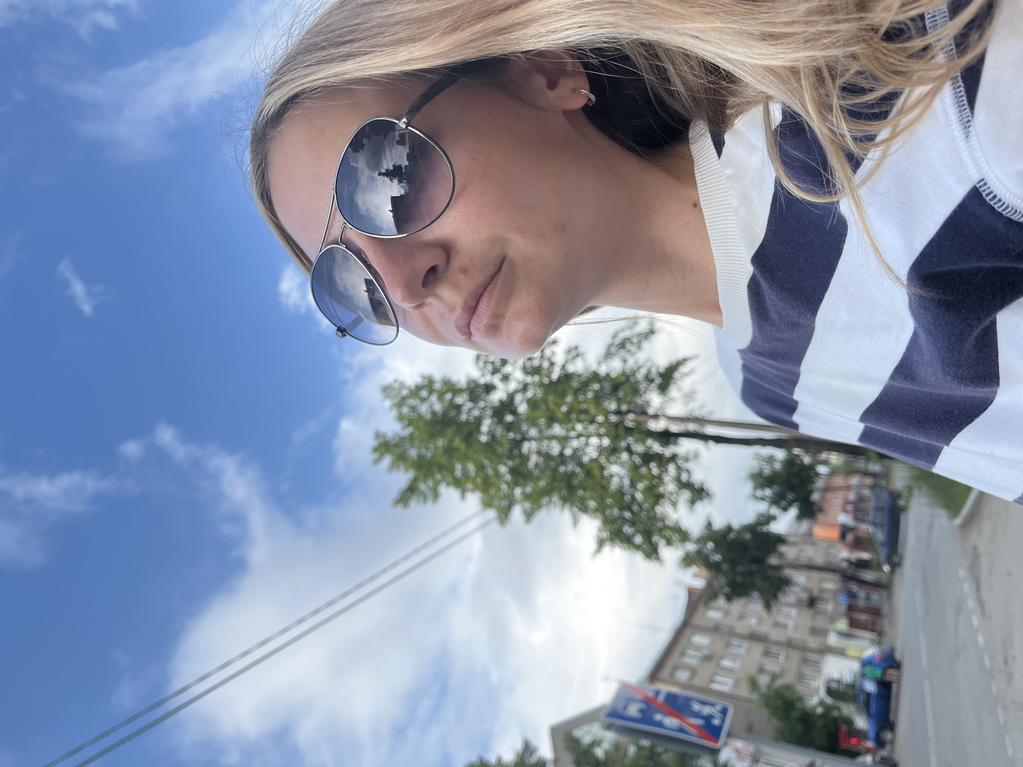 The image shows a young woman with blonde hair wearing aviator sunglasses and a navy and white striped long-sleeved shirt. She is looking upwards and to the right, with her face tilted towards the sky. The background is a city street scene in Chaikovsky, Russia, with buildings, trees, a street sign, and cars. The sky is blue with some white clouds, and power lines are visible across the top of the frame. The reflection in her sunglasses shows buildings and clouds.