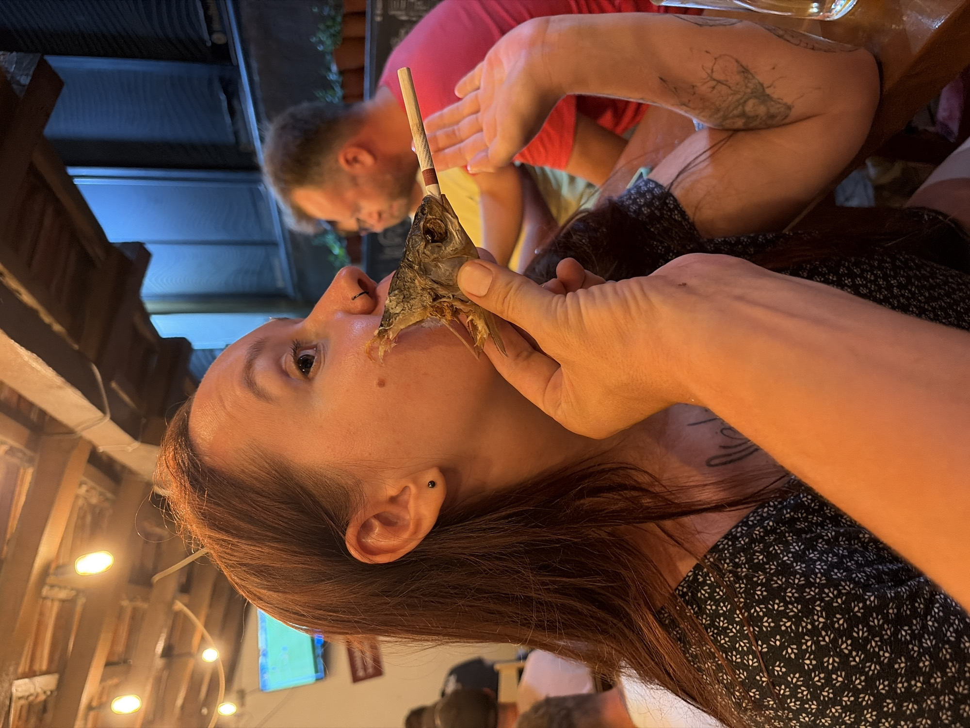 In Bar, Montenegro, a close-up shot captures a woman with long, reddish-brown hair, wearing a dark dress with a white floral pattern, as she holds the head of a fish to her mouth. The fish's mouth is open, revealing its skeletal structure, and a cigarette is inserted into it, protruding from its mouth. The woman has a black nose ring and a small black stud earring. Her left arm is visible, bent at the elbow, and a tattoo of a tree-like branch is visible on her forearm. Her right hand is extended, palm up. In the background, out of focus, a man wearing a red t-shirt is visible, as well as another man with short hair. The scene appears to be set indoors, with exposed wooden ceiling beams and string lights visible overhead. The lighting is warm, suggesting an evening setting.