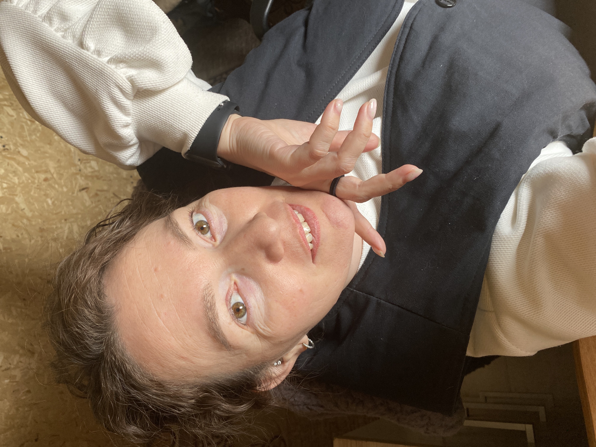 A woman is posing for a selfie, tilted at an angle, indoors. She has fair skin, light brown eyes, and curly brown hair with visible gray strands. She is smiling, showing her teeth. She is wearing a white textured sweater or sweatshirt under a black sleeveless jacket with a button visible. On her left wrist, she wears a black fitness tracker or smartwatch. She has a black ring on her left hand's ring finger. The background appears to be made of light-colored wood panels, possibly walls or furniture. There is no information to indicate this image was taken in Ol'khovskoye, Russia.