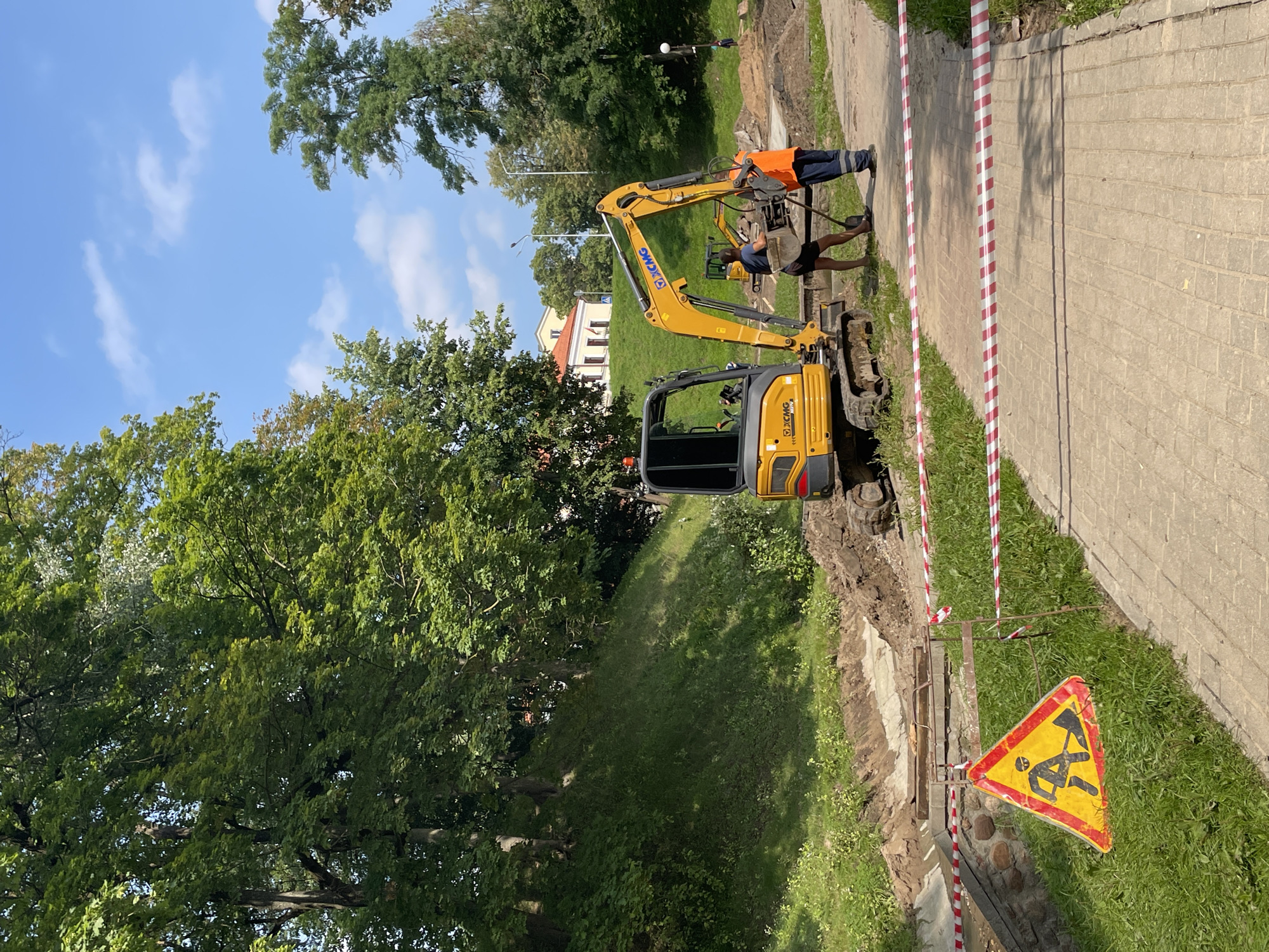The image depicts a construction scene in the city of Hrodna, Belarus. In the foreground, a yellow excavator is parked on a dirt path. Two construction workers are present near the excavator; one is wearing an orange vest and the other is in a dark t-shirt. They appear to be working with a shovel and a bucket. A red and white caution tape is strung across the path, indicating a restricted area. A yellow triangular sign with a black silhouette of a person digging is also visible on the left side of the image, reinforcing the construction warning. The background shows a grassy slope with trees and a building in the distance. The sky is blue with scattered white clouds. The overall scene suggests ongoing civil engineering work in an outdoor, possibly park-like, environment.
