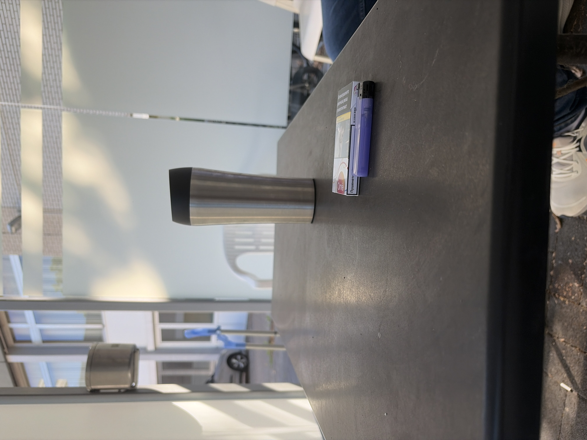 A stainless steel travel mug with a black lid sits on a dark, textured table outdoors. Next to the mug is a pack of "One" brand cigarettes and a purple disposable lighter. The table is positioned outside a building with large windows and frosted glass panels. In the background, a white plastic chair is visible, and a silver car is parked. The ground appears to be paved with square tiles. The scene is lit by natural daylight.