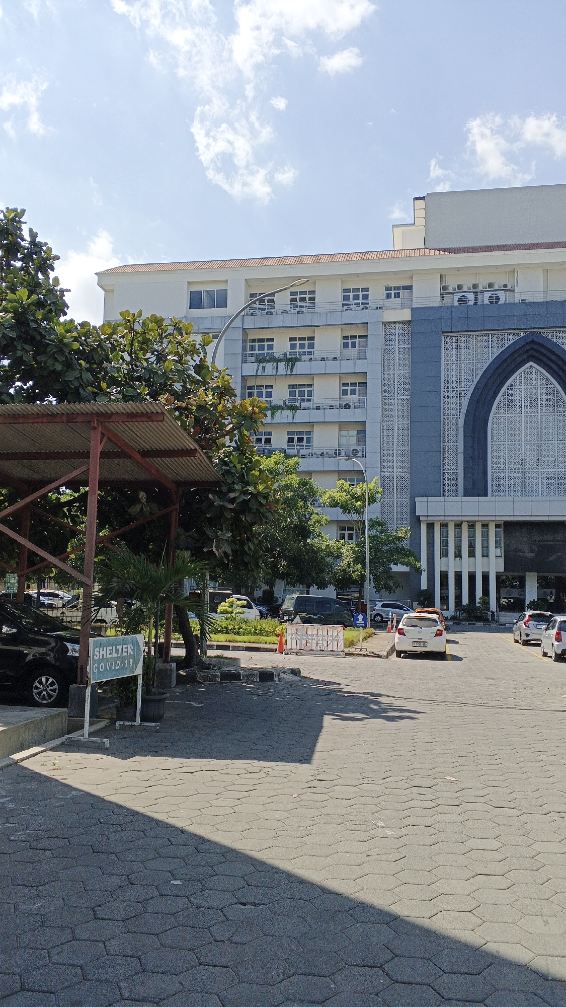 The image captures a sunny outdoor scene in Banguntapan, Indonesia, likely on the grounds of a large building such as a hospital or university. In the foreground, a paved area with hexagonal gray interlocking pavers is partially shaded by trees. To the left, a black car is parked near a covered structure with a corrugated metal roof, supported by red metal posts. A white sign with green text is visible under this structure, reading "SHELTER COVID-19".
Behind the shelter, lush green trees with broad leaves dominate the middle ground. A series of cars, including a white hatchback and other vehicles, are parked or driving on a paved road that curves towards a multi-story building in the background. This building is modern in design, with a pale exterior, multiple floors featuring numerous windows, and visible air conditioning units on some balconies. The right side of the building has an ornate, arched architectural feature with intricate white latticework.
The sky is bright blue with scattered white clouds, indicating fair weather. The overall impression is one of an active but orderly environment, with a clear focus on public health measures as indicated by the "SHELTER COVID-19" sign. The perspective suggests the photo was taken from a slightly elevated position, looking down into the parking area and towards the building.