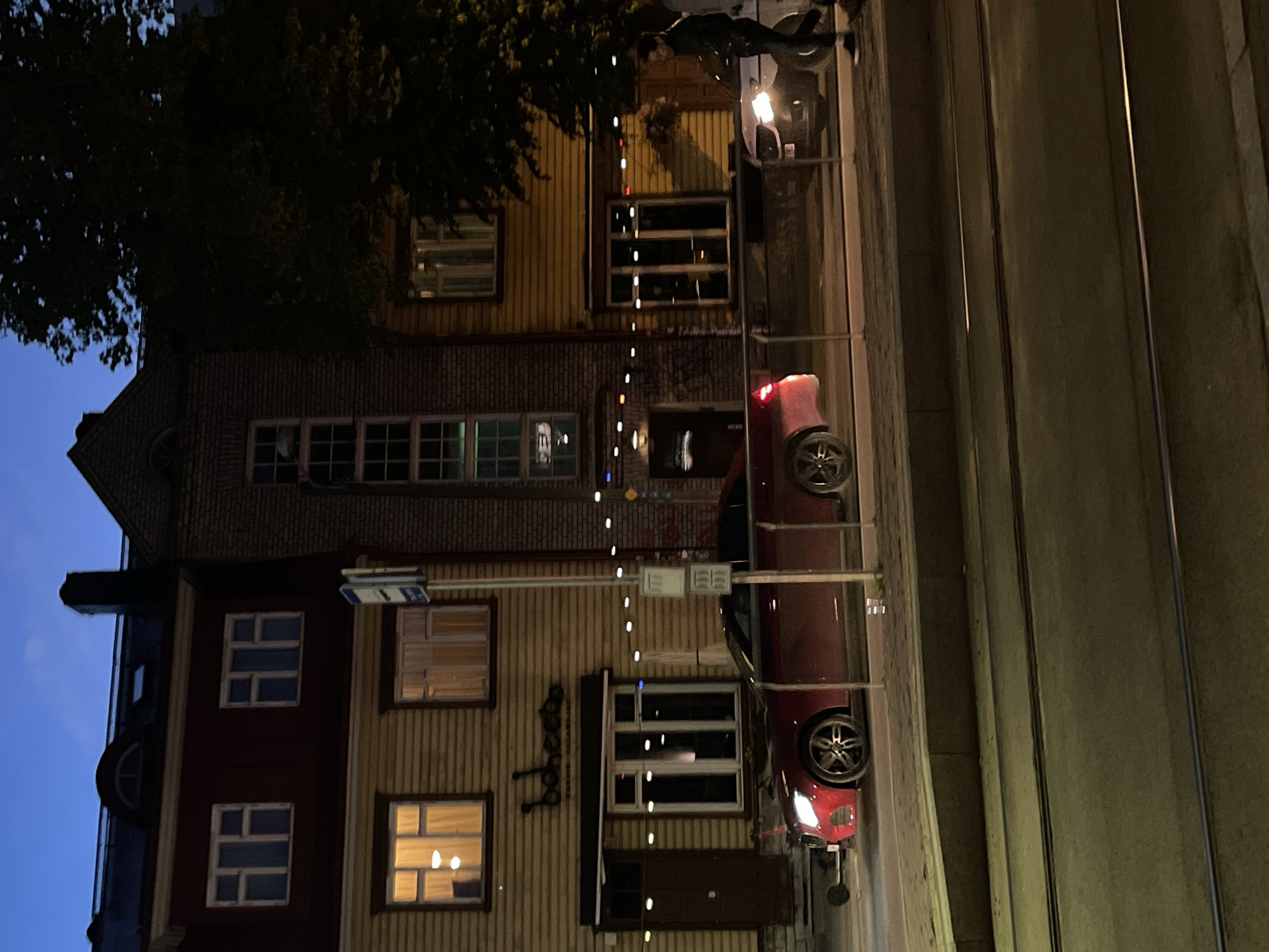 In Tallinn, Estonia, at dusk, a red Mercedes-Benz sedan is parked on the side of a street. The vehicle is positioned in front of a yellow wooden building with multiple windows and a dark red facade on the upper level. Above the building's entrance, a sign reads "babacoo". A person is walking away from the camera on the right side of the image, near a white BMW. The street in the foreground features tram tracks. The sky is a deep blue, indicating late evening.