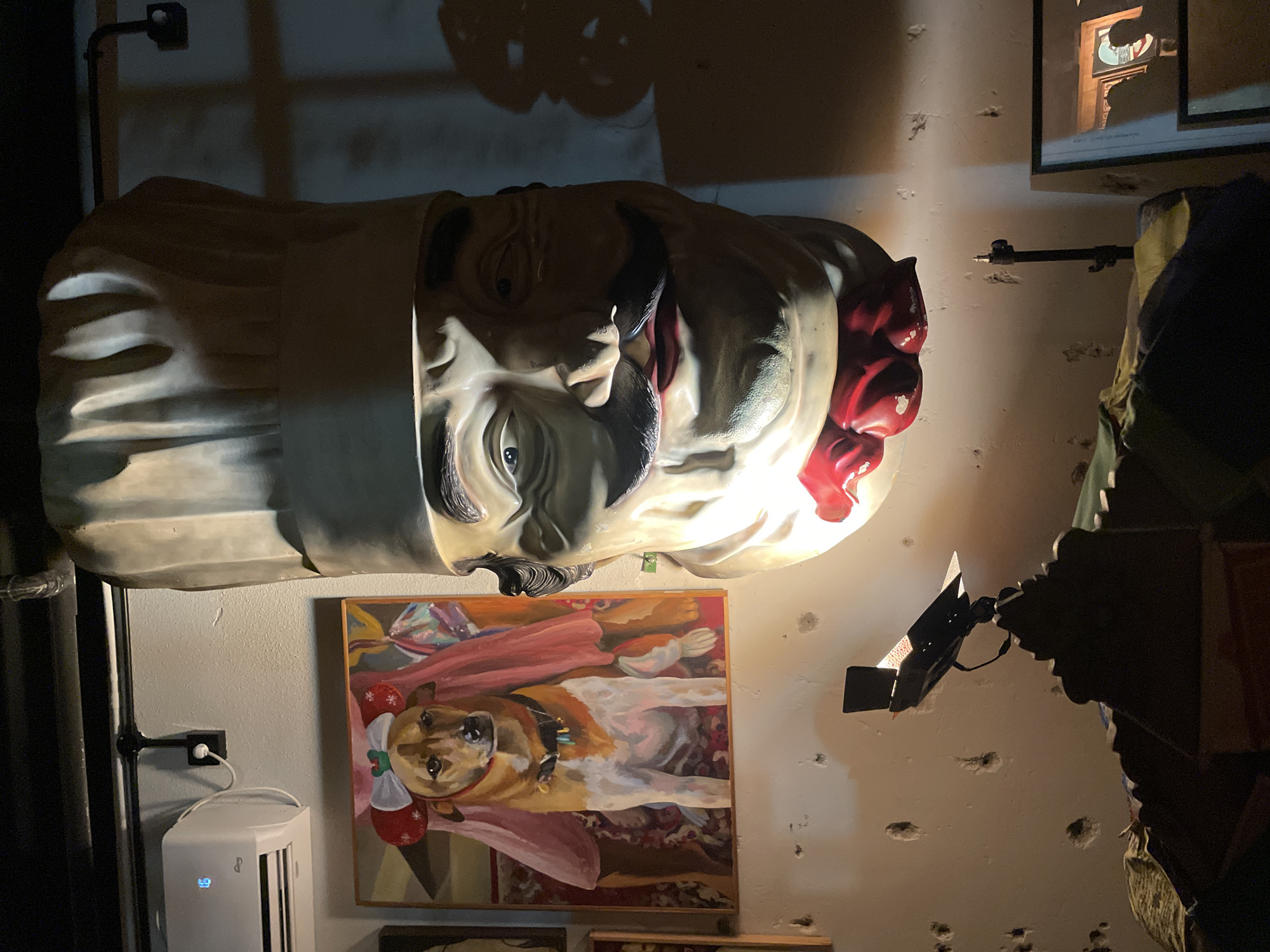 The image displays an artistic indoor scene, featuring a large, stylized bust of a man's head mounted on a light-colored, textured wall. The bust has prominent features including a dark mustache, dark hair, and intense eyes, with a white, pleated structure resembling a chef's hat around its base. A notable detail is the rear-right portion of the head, which appears to be "sliced" open, revealing intricate, red, brain-like material. Dramatic lighting from spotlights illuminates the bust and the wall, casting prominent shadows, including complex shapes above the bust.
Below the bust, a framed painting hangs on the wall. It depicts a light brown and white dog, possibly a Boxer or similar breed, lying on a patterned rug. The dog is adorned with festive red and white elements, resembling small mittens or bows, around its neck.
To the lower left of the painting, a white, rectangular electronic device, likely an air purifier or similar appliance, stands on the floor. Its digital display shows the number "69" in blue light. A small, stylized logo, possibly "LG" or similar, is visible below the display. A black pipe or conduit runs vertically along the left wall, connecting to an electrical outlet.
On the right side of the scene, a black spotlight on an adjustable arm is visible, directed towards the bust. Further right, a dark, jagged structure or display is partially visible. Above the spotlight, a framed picture hangs, but its contents are indistinct due to lighting and blur. The walls throughout the scene are a light color with a textured, uneven surface and numerous small indentations or holes. The overall atmosphere is artistic and somewhat surreal.
