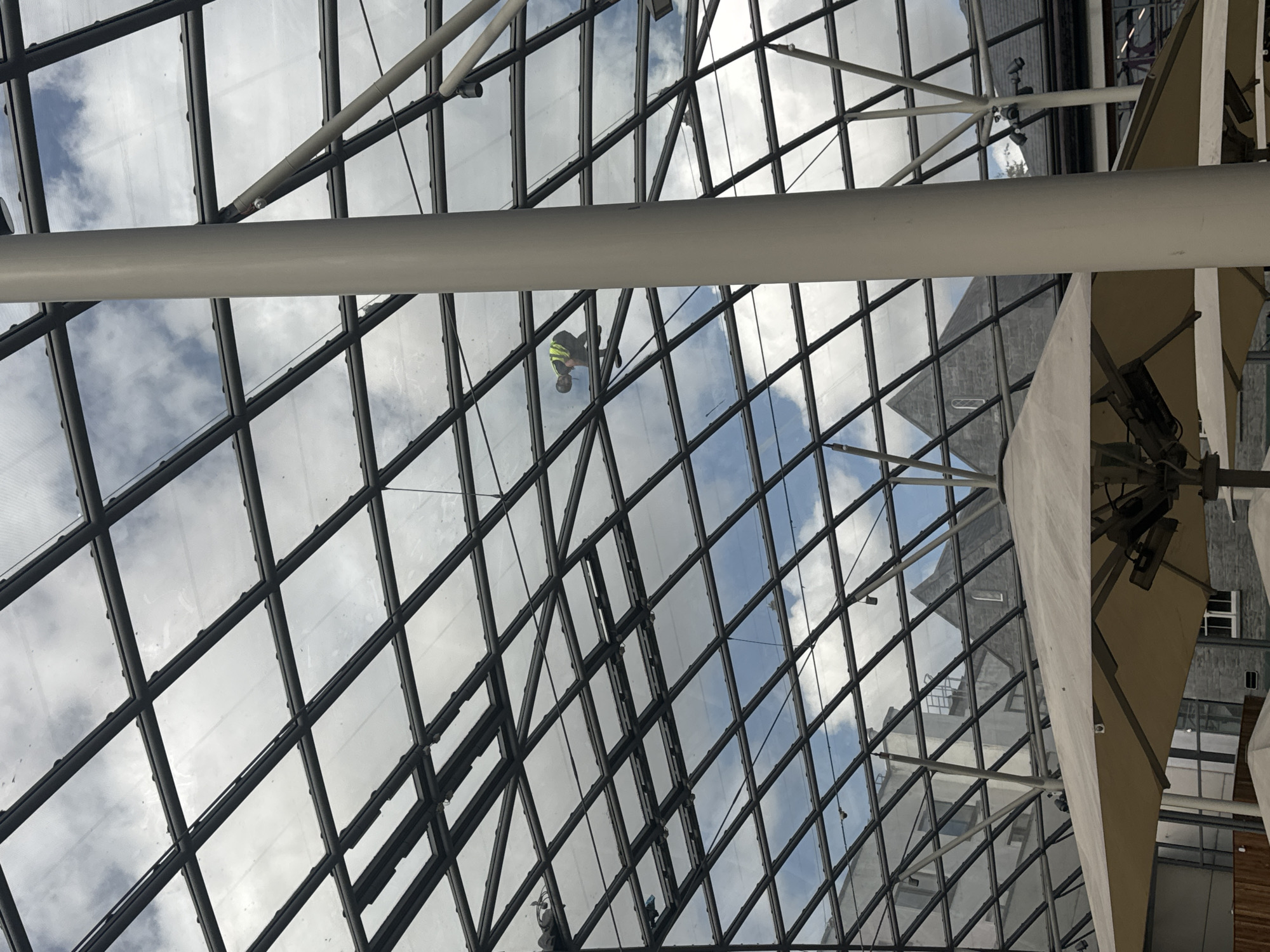 An individual wearing a yellow high-visibility vest and a hard hat is seen working on the interior side of a large, grid-like glass ceiling structure. The individual appears to be engaged in cleaning or maintenance. Below the glass ceiling, large cream-colored umbrellas are visible, suggesting an outdoor or semi-outdoor public space. In the background, the architecture of stone buildings with pitched roofs can be discerned through the glass, hinting at a historical or traditional urban setting. The sky, visible through the transparent ceiling panels, is partly cloudy. This scene is likely located within the Kilkenny Castle grounds in Kilkenny, Ireland, specifically in an area designed to cover a courtyard or an entrance.