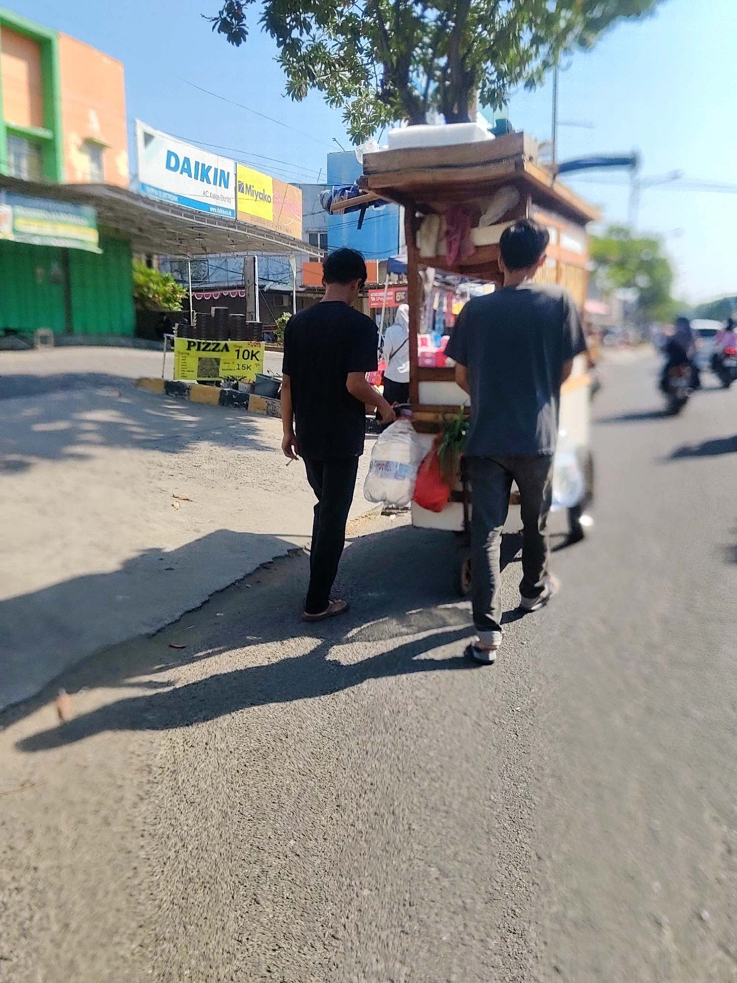 The image shows a street scene in Cibatu, Indonesia, on a sunny day. In the foreground, two men are walking on the right side of the road. The man on the left is wearing a black t-shirt and black pants. He is walking in the direction away from the camera. He is holding a clear plastic bag containing some items. The man on the right is wearing a dark gray t-shirt and dark gray pants. He is also walking away from the camera, pushing a cart. The cart has a wooden frame and a canopy. It contains various items, including what appears to be food and drinks. A red bag is visible on the cart.
In the background, there are buildings and trees. One building on the left has an orange and green facade, with a large sign that says "DAIKIN" and "Miyako." There is also a yellow sign on the left with "PIZZA 10K" written on it. Further down the street, there are other buildings and some people on motorcycles. The road is paved, and there are shadows cast by the people and the cart. The sky is clear and blue. The overall scene depicts a typical street view in a commercial area of Cibatu, Indonesia.