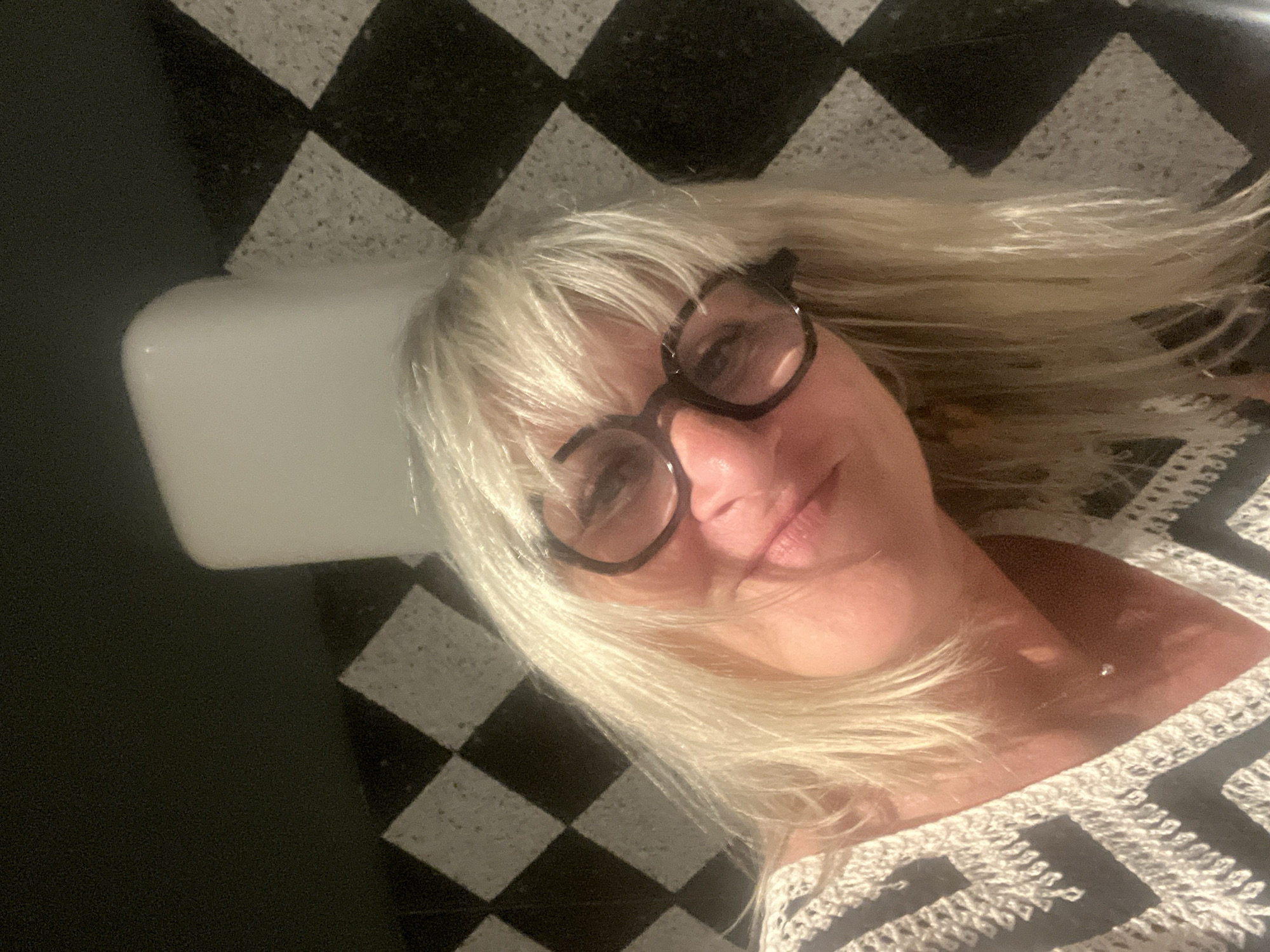The image shows a close-up, selfie-style portrait of a blonde-haired woman wearing dark-rimmed glasses and a patterned top. She is looking towards the camera with a slight smile. The background features a distinctive black and white checkered tile pattern on a wall, and a white, rectangular object, possibly a light fixture or part of a structure, is visible behind her. The overall impression is of an indoor setting with a modern or retro aesthetic. The image does not contain enough information to determine its specific location in the city of Antibes, France.