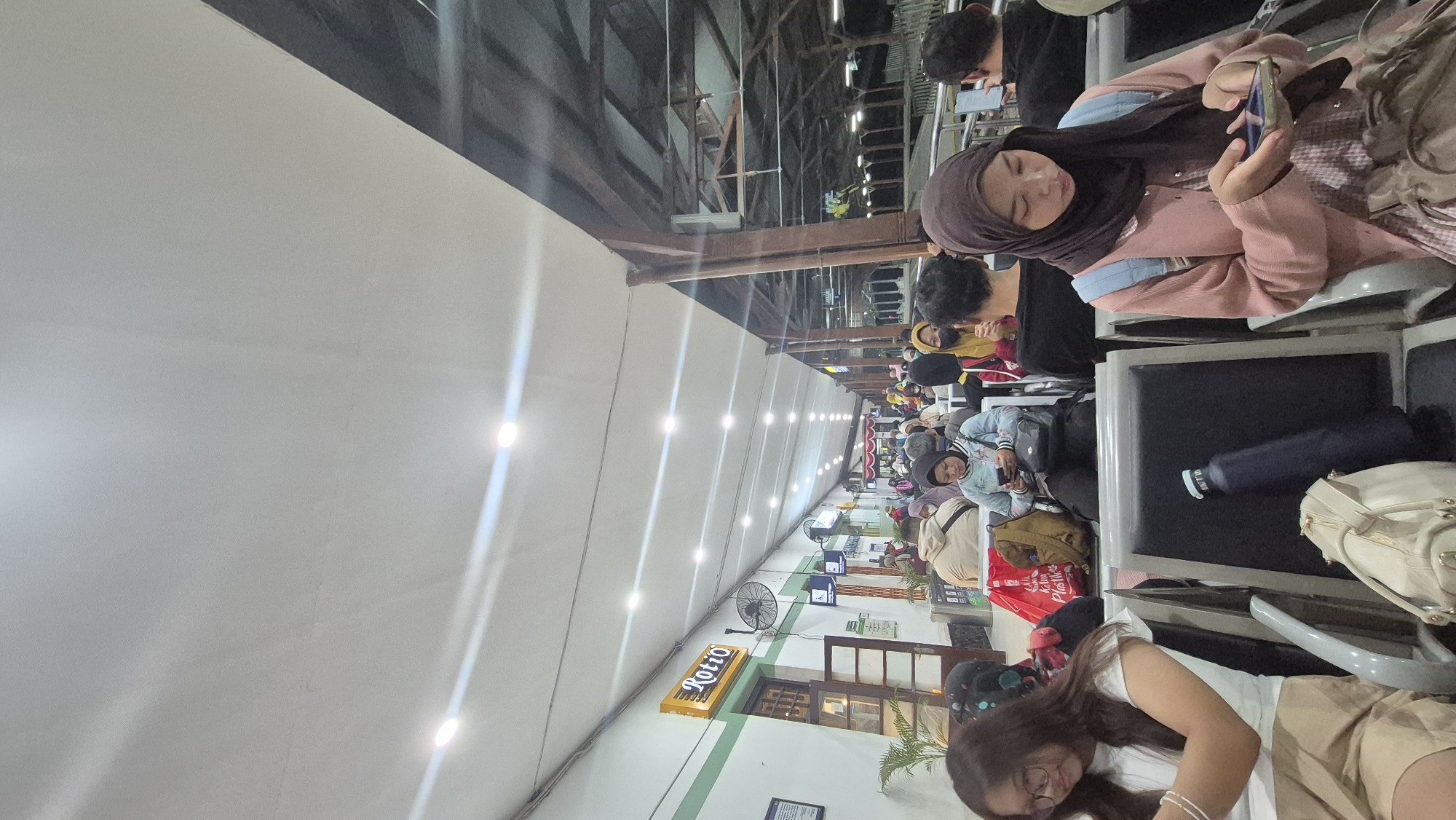 The image shows a crowded waiting area at the Bojonegoro train station in Bojonegoro, Indonesia. Numerous people are seated on benches, many of them looking at their phones. The environment appears to be a covered outdoor space, with a large white awning overhead and wooden support structures visible in the background. Fluorescent lights are mounted along the ceiling, illuminating the scene. To the left, there is a building with a sign that reads "Rotio" in an orange rectangle. Several plants are placed near the building. Bags and other personal belongings are scattered around the seated individuals. The overall impression is one of a busy transportation hub during a period of waiting.
