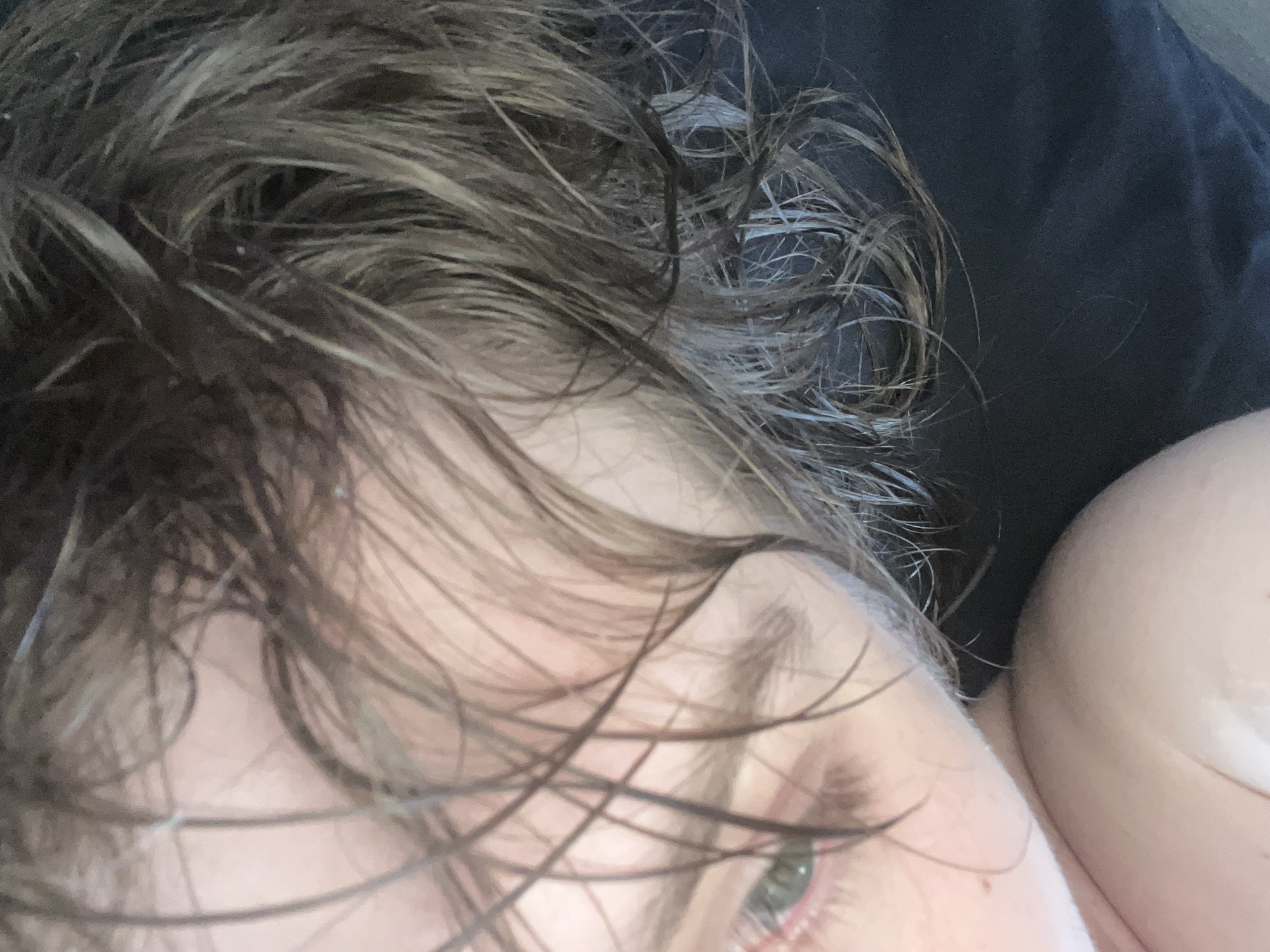 The image presents a close-up, top-down view of a person lying down in Saint Petersburg, Russia. The subject's head is tilted, displaying dark, disheveled hair that appears wet or moist, covering a significant portion of the left side of the frame and obscuring parts of the face. A section of the subject's face is visible on the right, featuring an open eye with a light-colored iris and prominent eyelashes, partially veiled by hair strands. The visible skin is fair-toned, exhibiting a reddish coloration around the eye area. In the upper right quadrant, a portion of a bare shoulder or upper chest is discernible. The background on the left and upper left consists of dark, solid-colored fabric, likely bedding or clothing. No specific actions or interactions are depicted beyond the subject's prone posture.