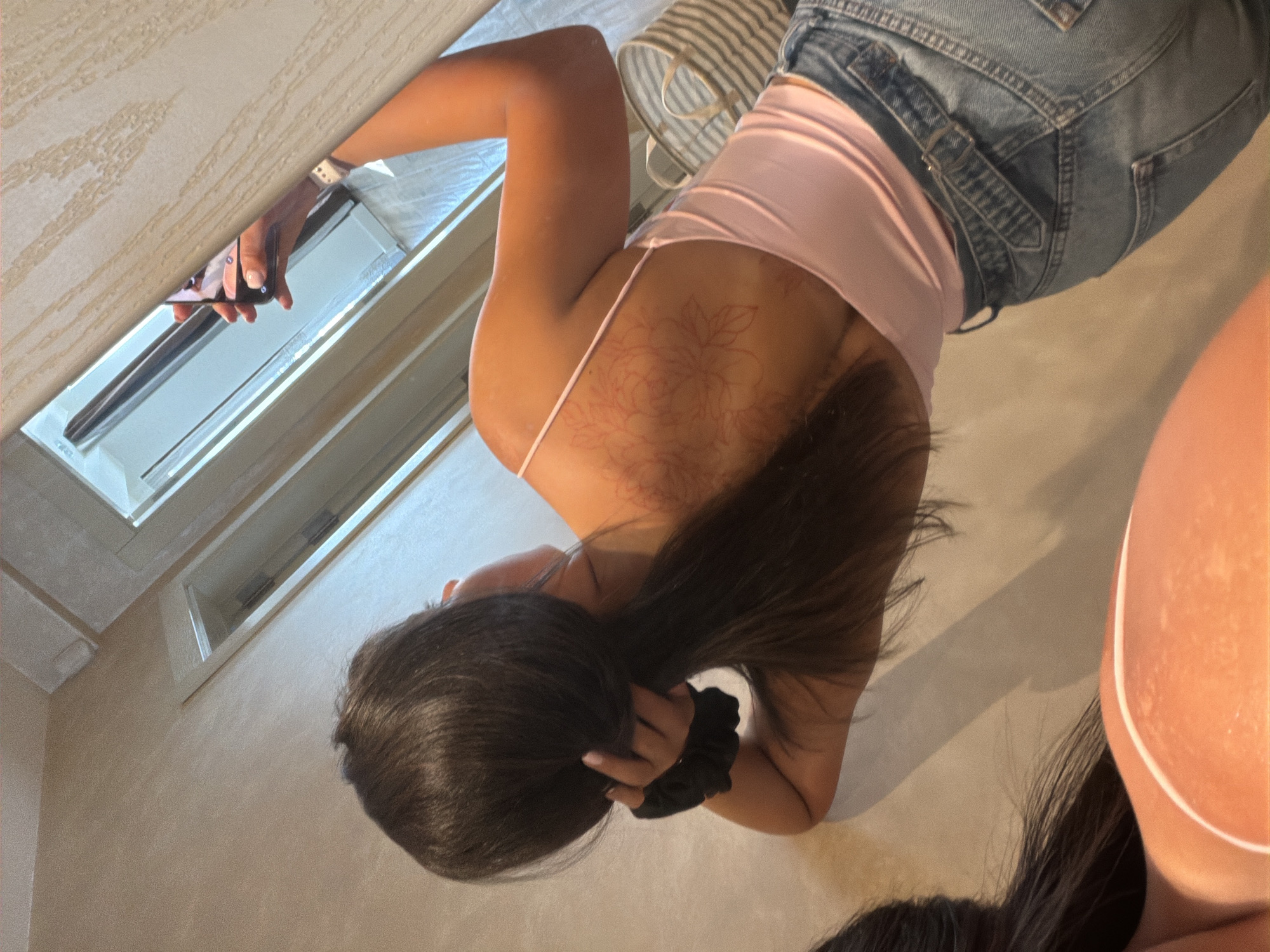 In the city of Gabrovo, Bulgaria, a young woman is captured in a mirrored selfie. She is wearing a pink spaghetti strap top and denim jeans. Her dark hair is pulled back into a ponytail. A faint floral tattoo is visible on her upper back. The background shows a lightly textured wall and what appears to be a doorway or alcove. A woven basket is partially visible on the right side of the image. The lighting is soft, suggesting an indoor setting.