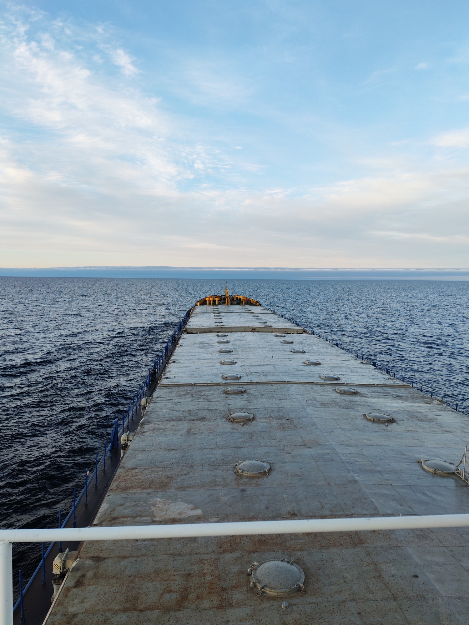 The image depicts a wide-angle view from the stern of a large vessel, likely a cargo ship or barge, looking forward towards its bow and the open water. The ship's broad, flat deck occupies the majority of the foreground and midground, appearing weathered and metallic gray, with numerous circular hatch covers evenly spaced across its surface. A prominent white handrail crosses the bottom of the frame, suggesting the photo was taken from an elevated position on the ship.
On either side of the deck, the deep blue sea stretches out, showing visible ripples and waves that indicate the vessel is in motion. Dark blue railings line the edges of the deck, separating it from the water. In the far distance, at the bow of the ship, some equipment and structures are faintly visible.
The upper portion of the image features a expansive sky, transitioning from a light blue overhead to a paler, more overcast tone near the horizon. Scattered, wispy white clouds are present throughout the sky. A distinct, horizontal band of darker, low-lying clouds or atmospheric haze forms a clear line where the sky meets the sea on the distant horizon.
The scene conveys a sense of peaceful transit across a vast body of water. The time of day appears to be daytime, with soft, diffused light, possibly early morning or late afternoon. There are no people visible in the image, nor is there any discernible text. The setting is open water, consistent with ocean or large sea navigation in the region of Unknown, Russia.