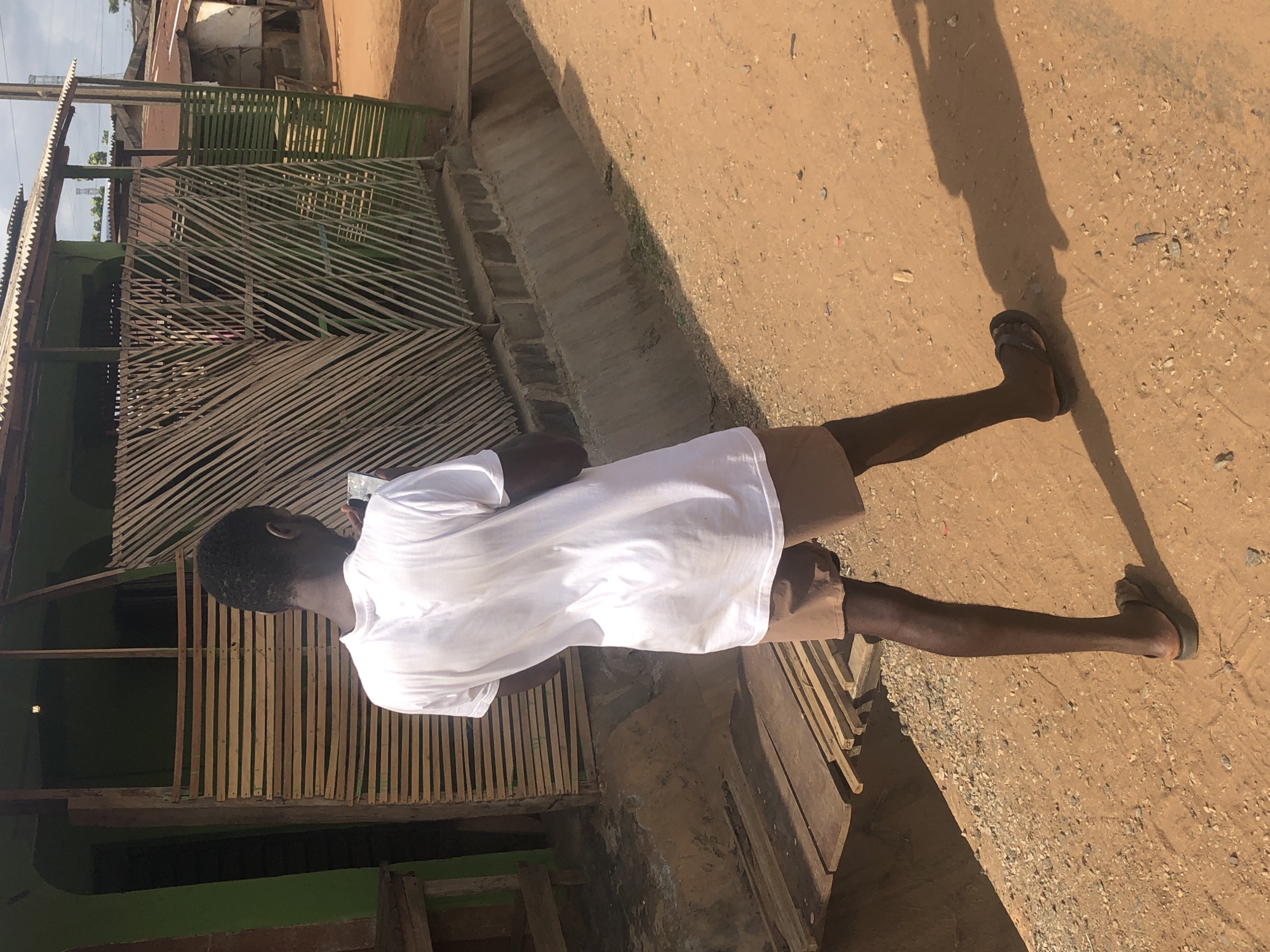 The image shows a person standing on a dirt road next to a building. The person is wearing a white t-shirt, brown shorts, and sandals. The building has a wooden lattice facade and a corrugated metal roof. The location is likely in a city with unpaved roads and simple structures. The city name is Unknown, Unknown.