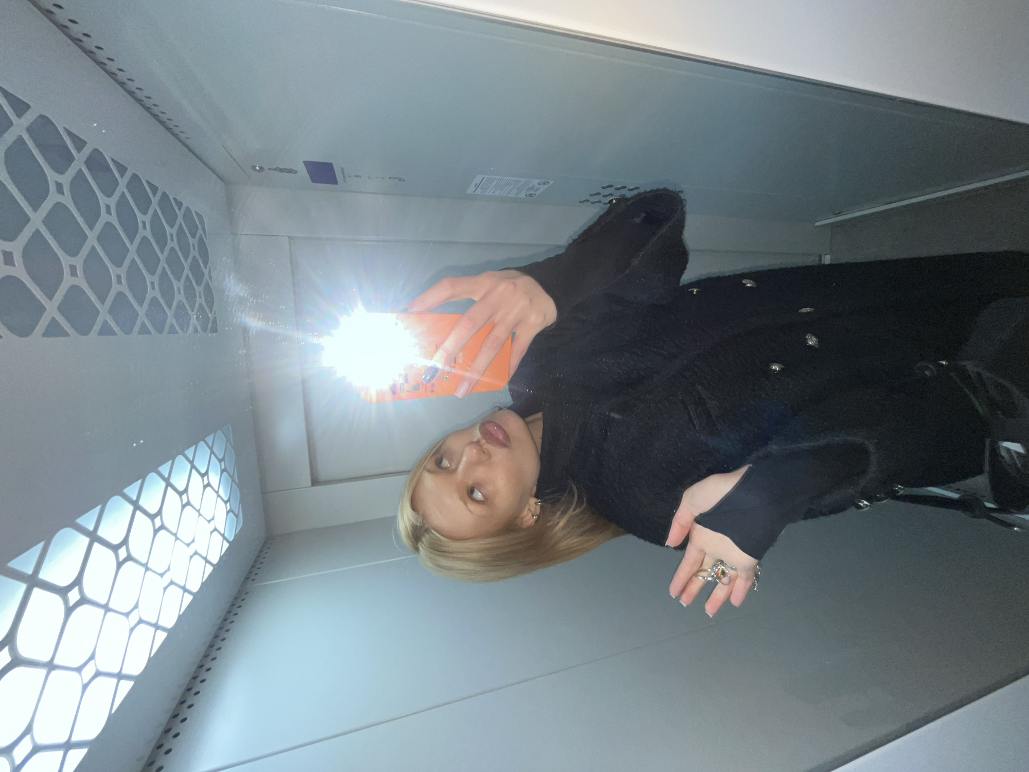 A blonde woman takes a mirror selfie inside an elevator in a building located in Chaikovsky, Russia. She is positioned centrally, gazing upwards with puckered lips and eyes rolled, holding an orange-cased smartphone in her right hand, which emits a bright flash that obscures part of her face. Her left hand is open, displaying multiple rings on her fingers. She wears a dark, textured coat with visible buttons over a black long-sleeved top. The elevator interior features light gray paneled walls with a vertical row of small circular perforations along the top edge. The ceiling is illuminated by a large rectangular light fixture with a white geometric grid pattern. A control panel with multiple circular buttons and a rectangular display is partially visible on the right wall.