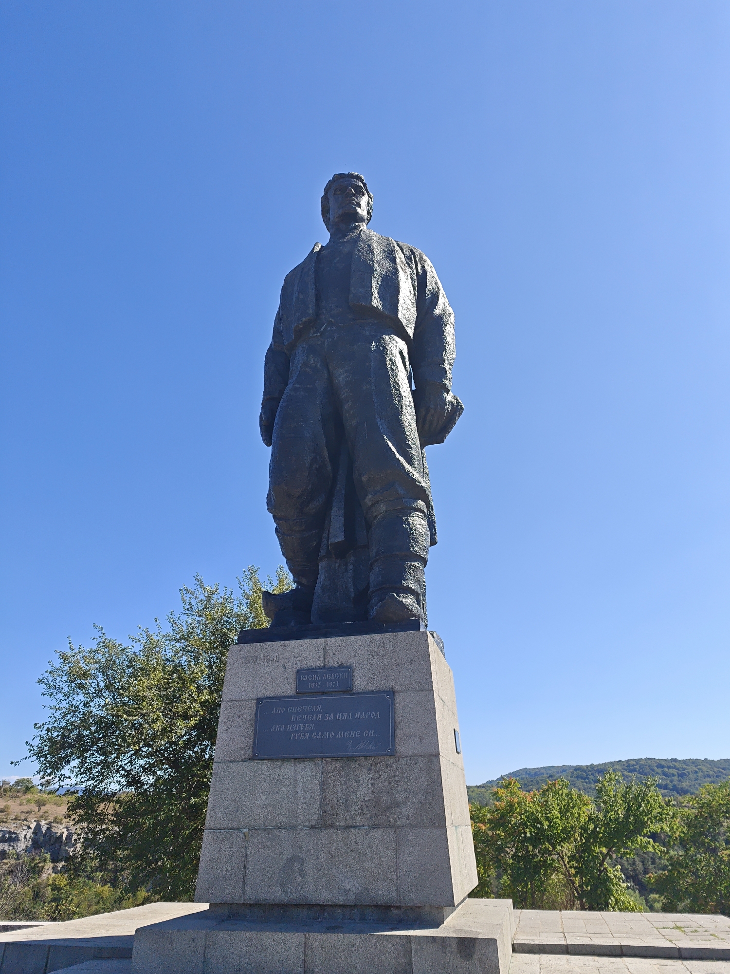 This is a photograph of a large bronze statue of a man, situated on a stone pedestal. The statue is dark in color and appears to be made of weathered metal, with visible texture. The man is depicted wearing traditional clothing, including a jacket and trousers, and is looking forward with a serious expression. The pedestal is made of grey stone blocks and features a plaque with writing. The background consists of a clear, bright blue sky, lush green trees, and rolling hills in the distance. The statue is located in Lovech, Bulgaria.