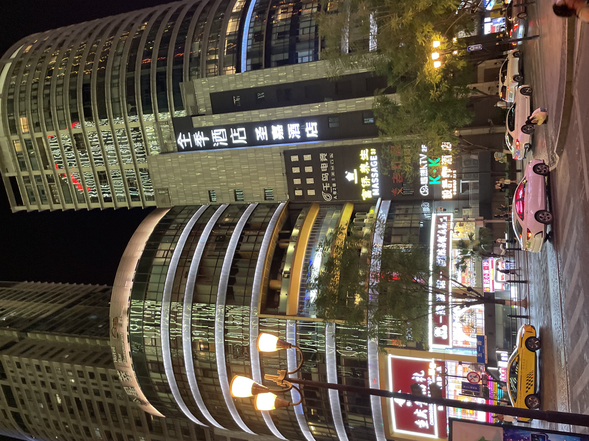 The image depicts a nighttime street scene in the Yuzhong District of China, characterized by illuminated modern architecture and urban infrastructure. In the foreground, several cars are visible on a wet asphalt road, including a yellow taxi and white sedans. Pedestrians are scattered on the sidewalks and crossing the street. Towering over the scene are multi-story buildings, one with a curved facade featuring prominent horizontal bands of white light. Another taller, rectangular building displays reflective windows that mirror the city lights. Signs with Chinese characters and English text, advertising businesses such as "Hotel Bar Restaurant," "MASSAGE 3F," and "KTV 2F," are illuminated on the lower levels of the buildings. A streetlamp with three globes stands on the left side of the frame, casting a warm glow. The environment suggests a busy commercial and residential area.