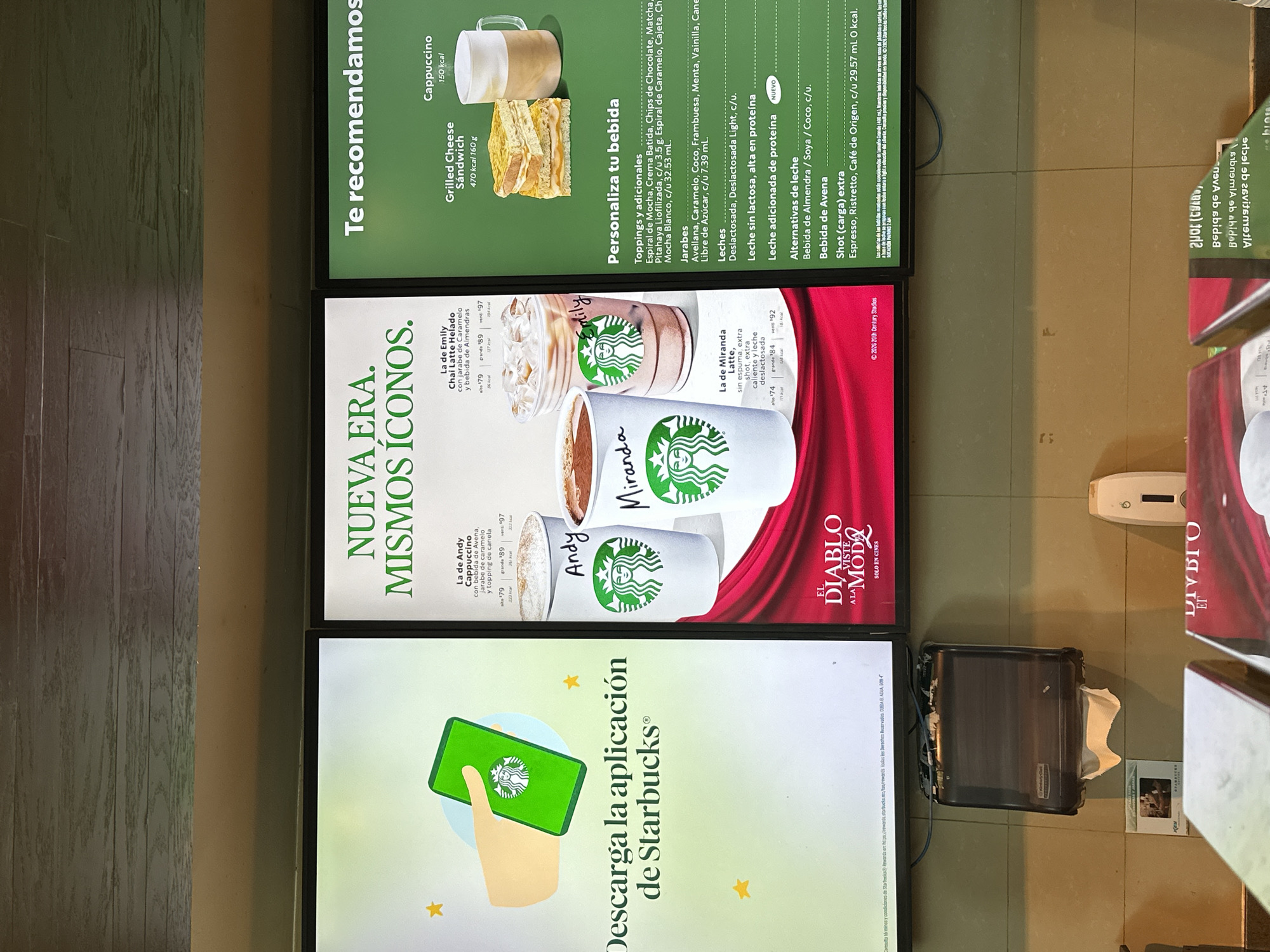 Three vertical digital screens, likely part of an indoor commercial setting in San Andrés Cholula, Mexico, display advertisements. The screens are bordered by black frames and mounted against a wall. To the left, a light wooden panel is visible, while to the right, a neutral-colored floor or wall can be seen, along with a black dispenser containing a white tissue, and a small white box.
The top screen is a Starbucks menu board titled "Te recomendamos" (We recommend), featuring a "Grilled Cheese Sandwich" (470 kcal 160g) and a "Cappuccino" (150 kcal). Below this, "Personaliza tu bebida" (Personalize your drink) offers various "Toppings y adicionales" (e.g., Mocha spiral, whipped cream), "Jarabes" (Syrups like Hazelnut, Caramel), "Leches" (Milks including lactose-free and high-protein), and "Alternativas de leche" (Milk alternatives like Almond, Soy, Oat). Prices and calorie counts are listed for many items.
The middle screen showcases "NUEVA ERA. MISMOS ÍCONOS." (NEW ERA. SAME ICONS.), depicting three personalized Starbucks drinks: "La de Emily" (Chai Latte Helado with Caramel syrup and Almond milk), "La de Andy" (Cappuccino with Oat beverage, Caramel syrup, and cinnamon topping), and "La de Miranda" (Latte, sugar-free, extra shot, extra hot, lactose-free milk). Each drink shows prices for different sizes (alto, grande, venti) and calorie information. Notably, this screen also features a movie advertisement for "EL DIABLO VISTE A LA MODA SOLO EN CINES" (The Devil Wears Prada, Only in Cinemas) with a visible copyright from "2026 20th Century Studios."
The bottom screen encourages users to "Descarga la aplicación de Starbucks®" (Download the Starbucks® app), illustrated by a hand holding a smartphone displaying the Starbucks logo. Small yellow stars are scattered across this screen. Partially visible text on the bottom right of the middle screen indicates "©2026 Starbucks Coffee Company."