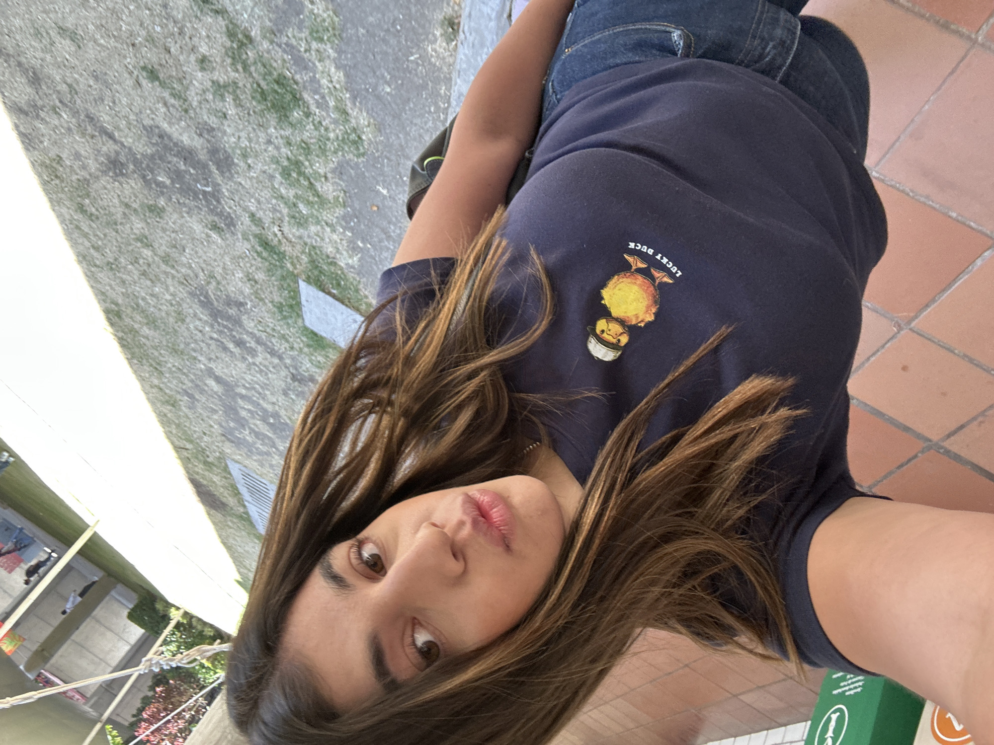 A young female, appearing to be of Latin American descent, is depicted in a high-angle selfie shot outdoors in San Bernardino Tlaxcalancingo, Mexico. She has long, straight, brown hair with lighter highlights, brown eyes, and her lips are slightly pursed. She is wearing a dark blue short-sleeved t-shirt with a yellow duck graphic wearing a hat, accompanied by the text "LUCKY DUCK." Blue denim shorts are visible from the waist down, and a dark backpack strap is partially visible over her right shoulder. The foreground consists of terra cotta-colored square floor tiles. To the lower left, a green object with white markings is partially visible. The background reveals an outdoor paved area transitioning into a patch of light green grass, with a metal grate present on the paved surface. Beyond this, an elevated open-air structure or walkway supported by pillars is visible, with multiple indistinct human figures present on it in the distance. Sparse green and reddish foliage is visible to the left of the background structure. The scene is illuminated by daylight, casting shadows on the ground.