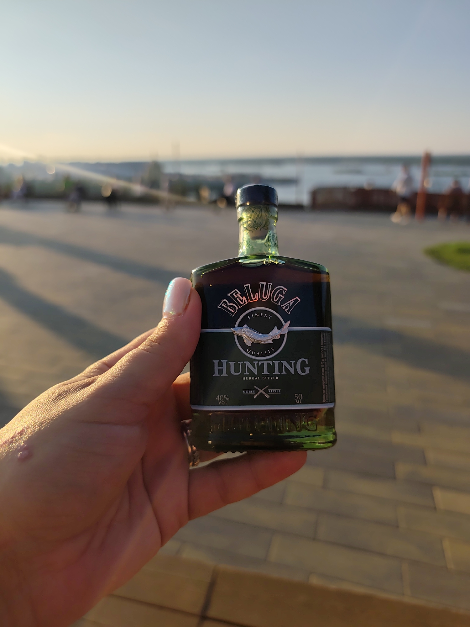 A person's hand holds a small, green glass bottle of Beluga Hunting Herbal Bitter in the foreground. The bottle has a black and green label with white text and a silver fish emblem. The background is out of focus and shows a body of water, possibly the Volga River, and a boardwalk or promenade with people and some streetlights. The setting appears to be a public outdoor space in Nizhny Novgorod, Russia, during sunset, indicated by the warm, golden light and long shadows.