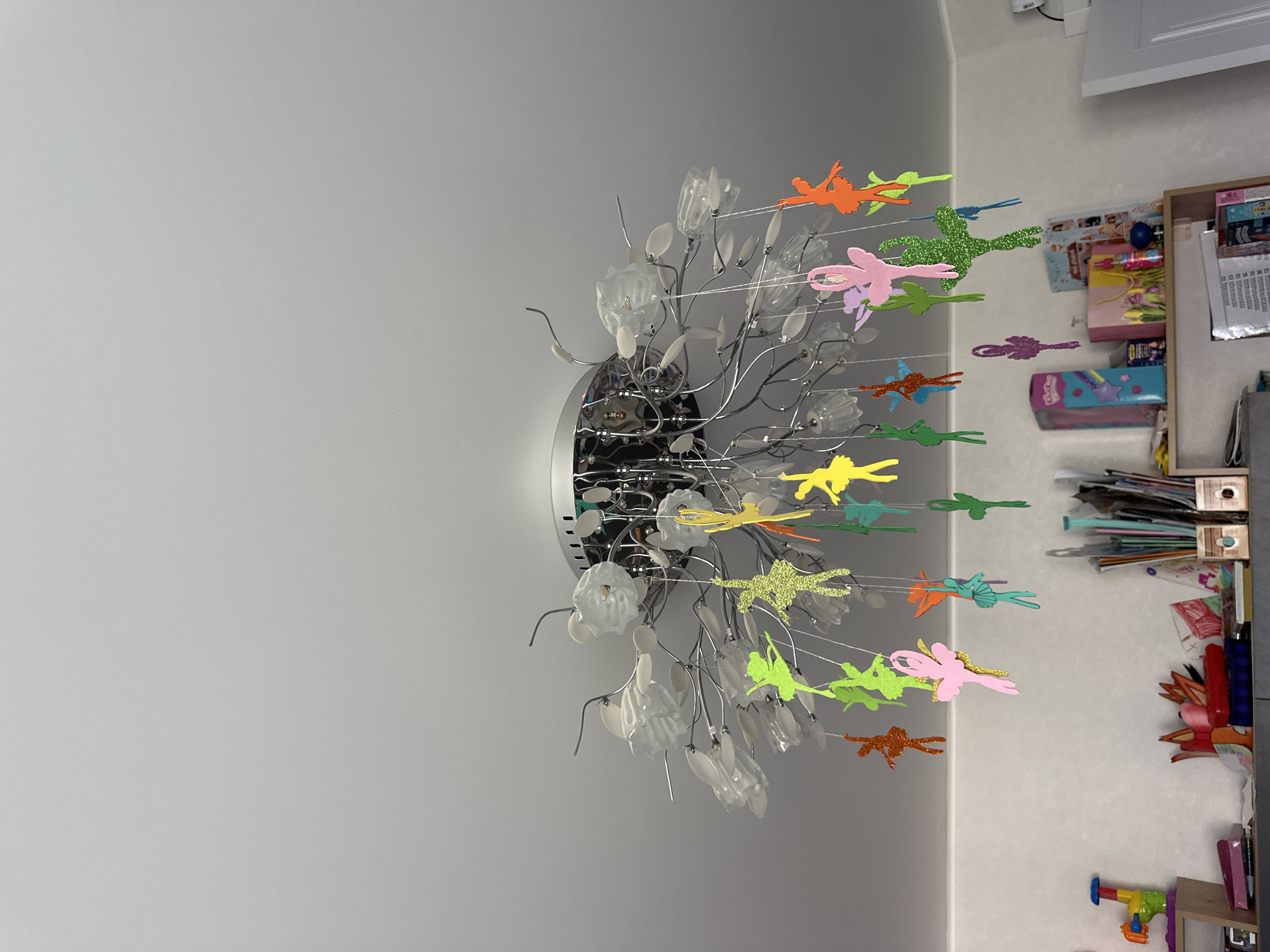 A close-up, eye-level shot shows a chandelier with multiple hanging glittered ballet dancer ornaments. The chandelier has a metallic base and decorative white flower-like accents. The dancers are in various poses and are colored pink, green, orange, yellow, and teal. The background consists of a white ceiling and a shelf unit with various items. The overall scene is indoors, likely in a room. The image does not contain any specific geographical markers to indicate its location in Мариинский, Russia.