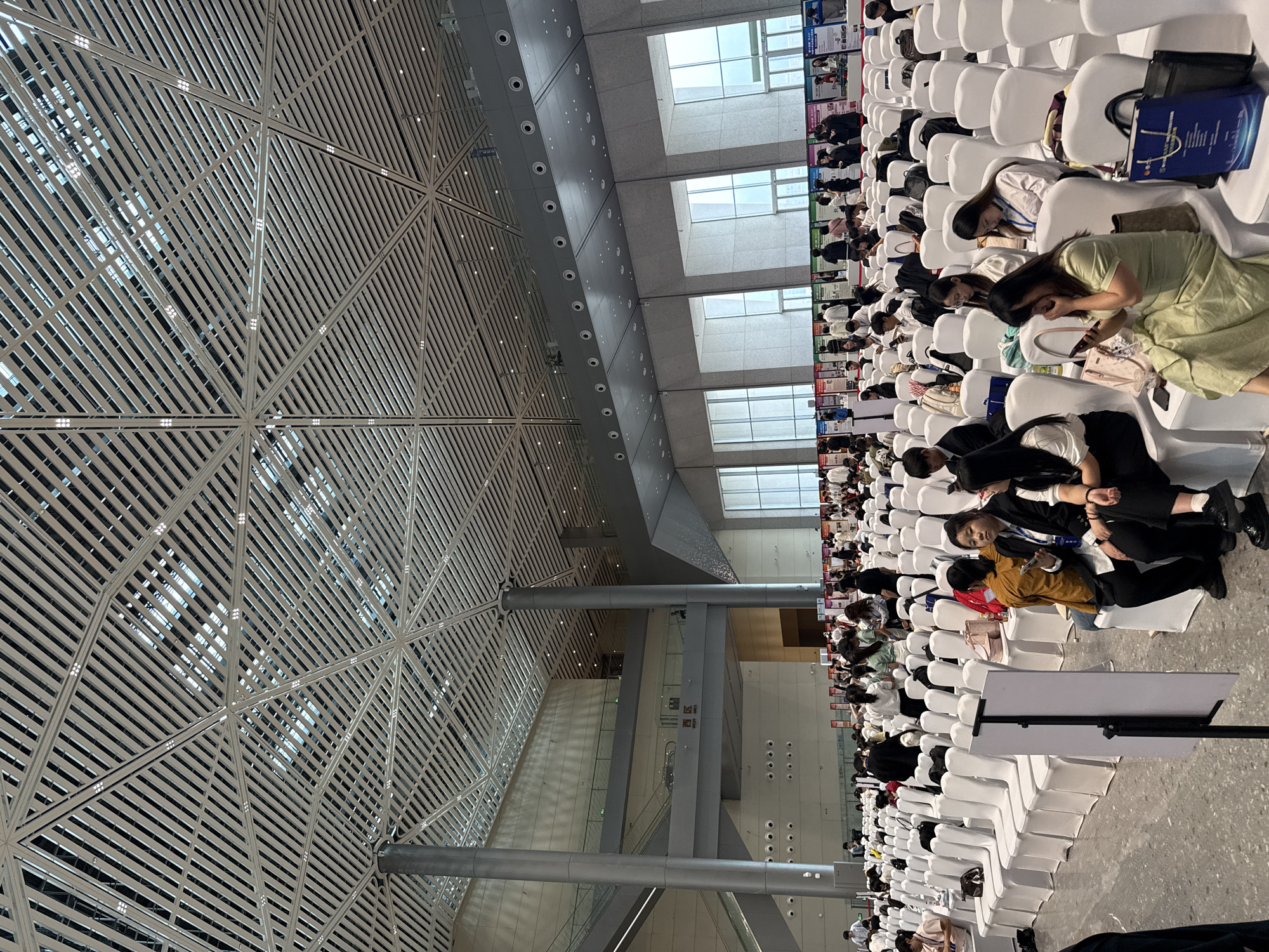 The image depicts a large indoor event space, likely a convention center or exhibition hall, in the city of Chongqing High-tech Zone, China. The venue features a modern, geometrically patterned ceiling with a complex network of beams and lights. In the foreground, rows of white chairs are arranged facing forward, indicating a seating arrangement for an audience or attendees. Numerous people, appearing to be young adults, are seated or standing in the space. Many are looking at their phones or interacting with each other. Along the back wall, there are several displays or booths set up, suggesting an exhibition or presentation area. The overall impression is one of a bustling event or gathering.