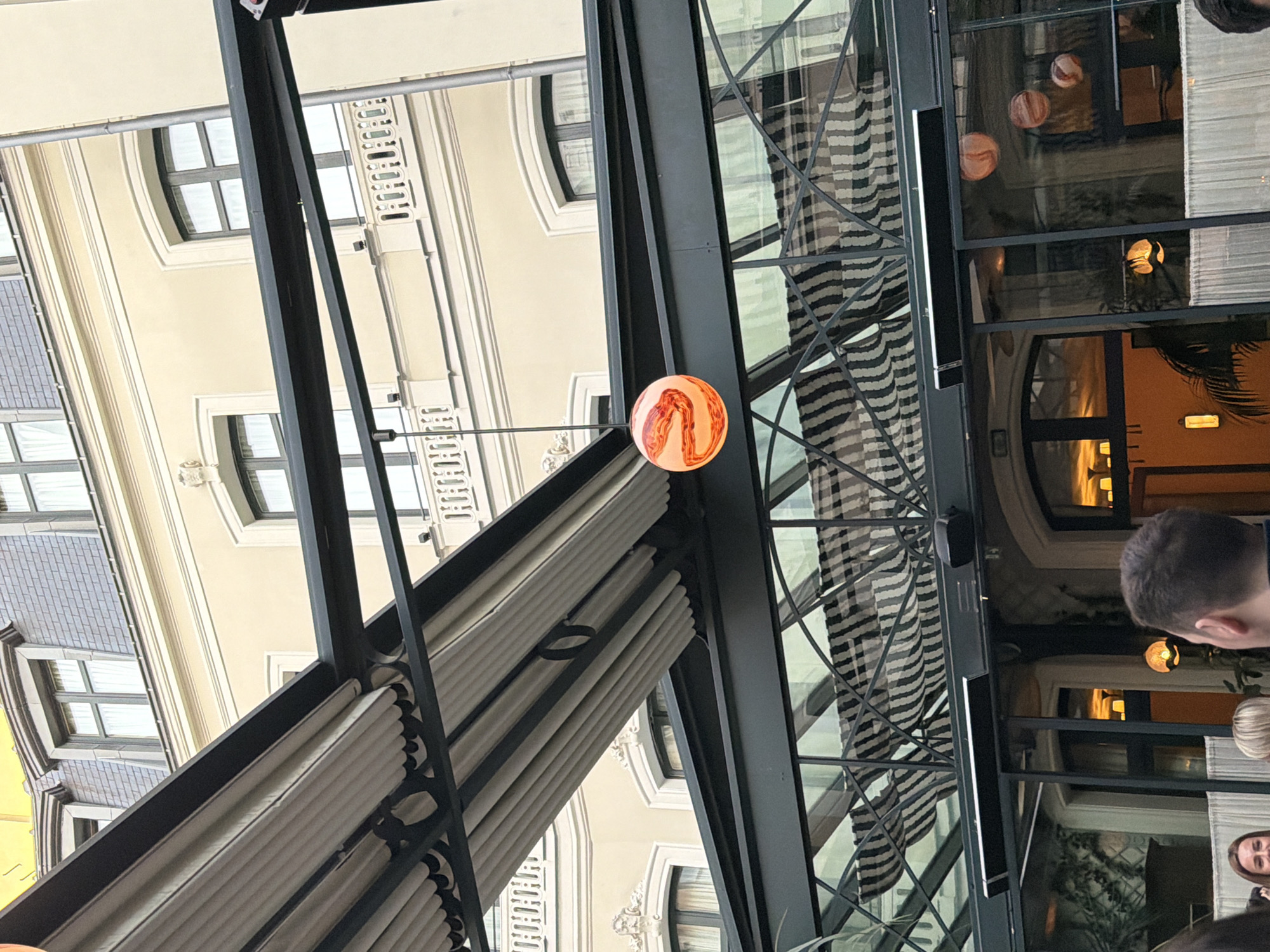 The image displays an upward view from a covered outdoor area, capturing reflections and architectural details, rotated 90 degrees clockwise from its natural orientation. A prominent black metal framework, likely part of a canopy or roof structure, dominates the scene. Hanging from this structure is a vibrant spherical light fixture, featuring orange and white swirling patterns, resembling a marbled or molten glass design. Below the main frame, light-colored retractable awnings or shades are neatly folded.
Through and reflected in the glass panels of the overhead structure, elements of both the interior space and the exterior buildings are visible. Reflections include dark and light striped patterns, possibly from curtains or upholstery, and several other golden-toned spherical lights hanging inside the establishment.
In the background, visible through openings in the structure and reflected in the glass, is the façade of a classic European building. It features light-colored, possibly cream or beige, stucco walls, arched windows with dark frames, and decorative architectural details, consistent with Parisian architecture. Another building section on the far left shows a darker, possibly tiled, upper story.
In the lower right corner, parts of people are visible, including the back of a man's head with short, dark hair, and a portion of a woman's face and blonde hair. Their presence suggests the setting is a café, restaurant, or a similar public outdoor area. The interior lights being on suggests it could be evening or an overcast day. No discernible text is visible in the image.