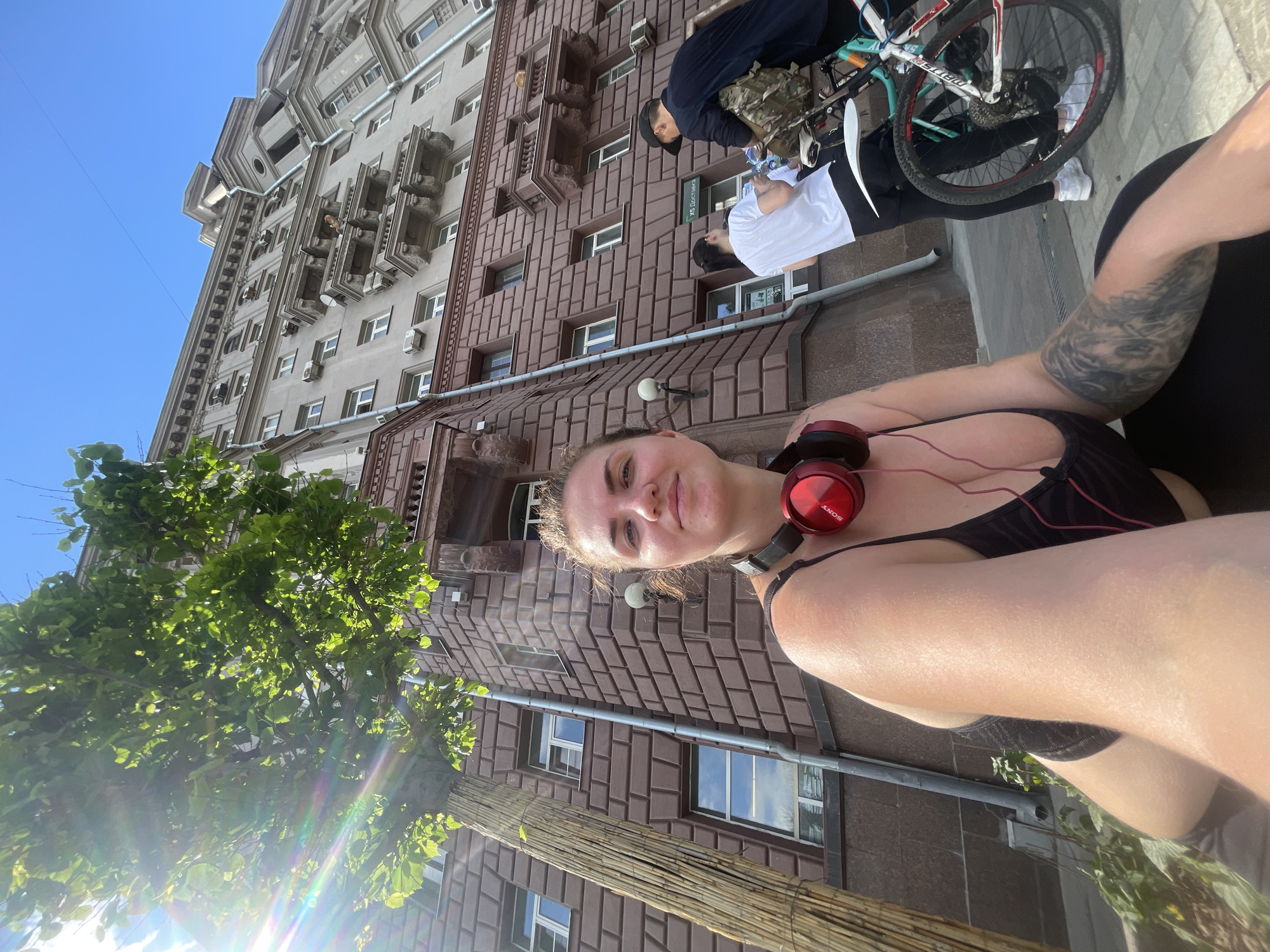 The image captures a low-angle perspective street view in Moscow, Russia. A woman wearing a black top and red Sony headphones is centered in the foreground, holding the camera for a selfie. She has a tattoo on her arm. Behind her is a multi-story building with a reddish-brown facade featuring geometric brickwork. To the left, a tree with green leaves is visible. A glimpse of a light gray building with ornate architectural details can be seen in the background. Two people are standing near a bicycle on the sidewalk near the reddish-brown building. One person is wearing a dark jacket and a baseball cap, while the other is wearing a white t-shirt.