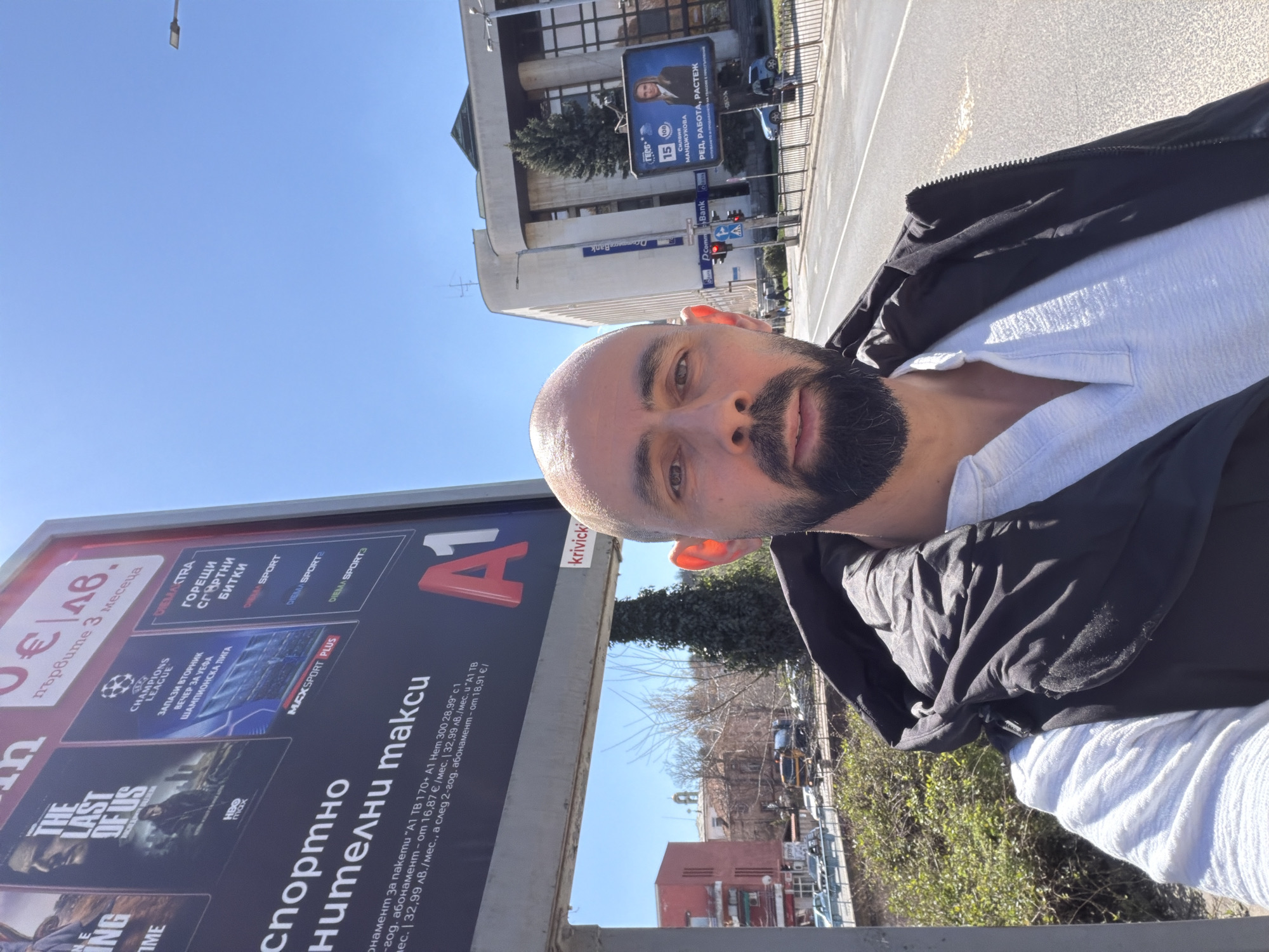 A selfie taken in Haskovo, Bulgaria, features a bald man with a beard wearing a white shirt and black jacket, standing next to a large A1 billboard advertising telecommunications services and HBO's "The Last of Us." The billboard also promotes Champions League soccer coverage. In the background, there is a street with buildings and cars, including what appears to be the D Commerce Bank, identifiable by its sign, as well as an advertisement featuring a woman with the text "GERB," followed by text about “RED, RABOTA, RASTEZH,” which translates to order, work, growth. The sky is blue and the sun is shining.