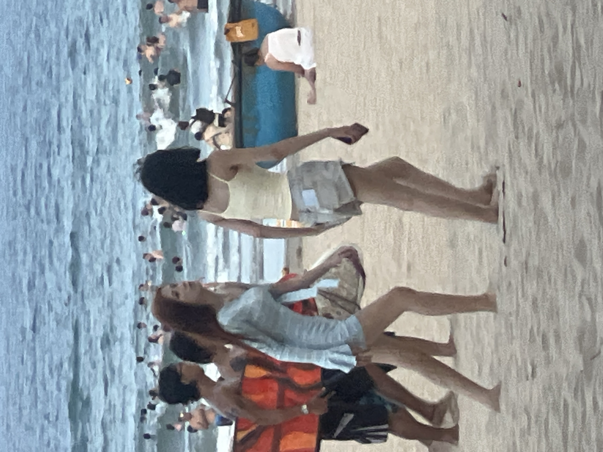 The image depicts a beach scene in Đà Nẵng, Vietnam, with several people enjoying the water and sand. In the foreground, three individuals are walking on the beach. A man on the left is carrying a surfboard with an orange and red abstract pattern. To his right, a woman with long auburn hair is walking in a light blue dress. Behind her, another woman with dark hair is wearing a yellow tank top and light-colored shorts.
In the background, the ocean is populated with numerous swimmers and bathers. A blue boat is visible near the shore, and a woman in a white top is sitting on the sand nearby. The overall atmosphere suggests a recreational day at the beach.